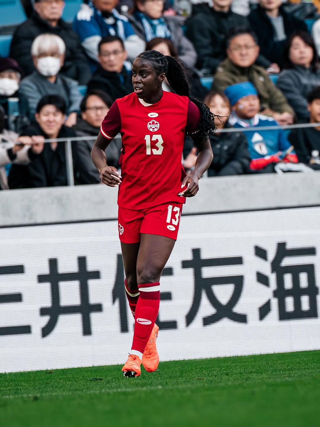 Simi Awujo #13 of Canada Women’s National Team in a red kit. The Manchester United midfielder of Nigerian heritage is shown on the pitch in orange cleats.
