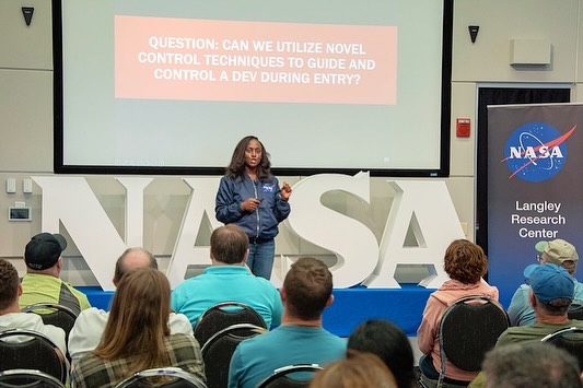 Dr Wendy Okolo presenting at NASA Langley Research Center in a NASA jacket, standing in front of a screen displaying a question about novel control techniques for space vehicles.