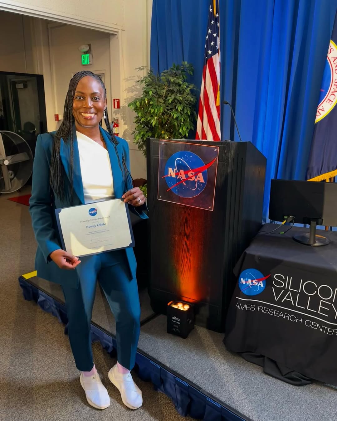 Dr Wendy Okolo smiling and holding her first patent certificate at the NASA Ames Technology Transfer award ceremony, wearing a teal suit and white trainers.