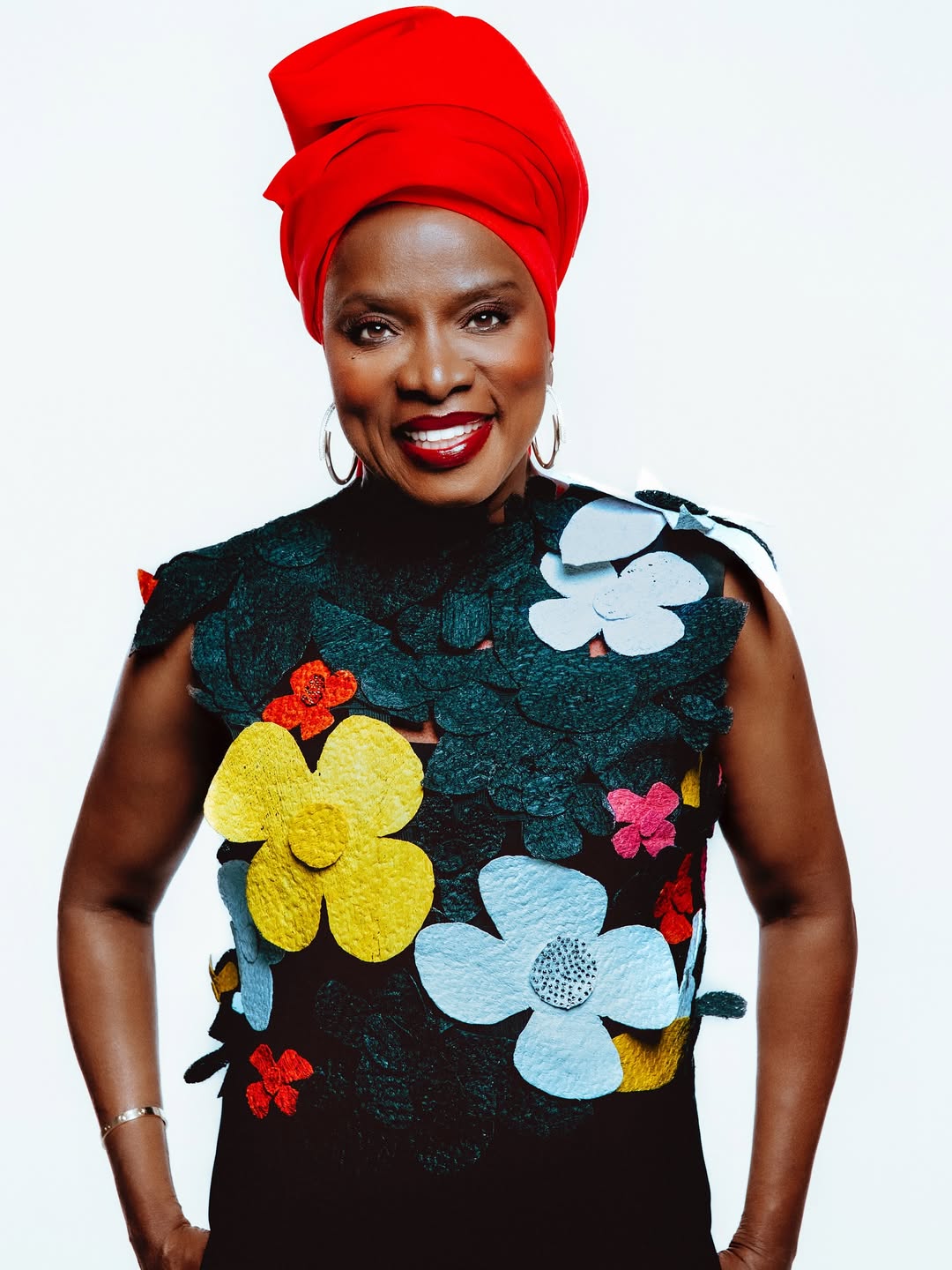 Close-up studio portrait of Angélique Kidjo wearing a red headscarf and a black top with colourful 3D floral appliqués, smiling against a white background for her "Hope!!" album campaign.