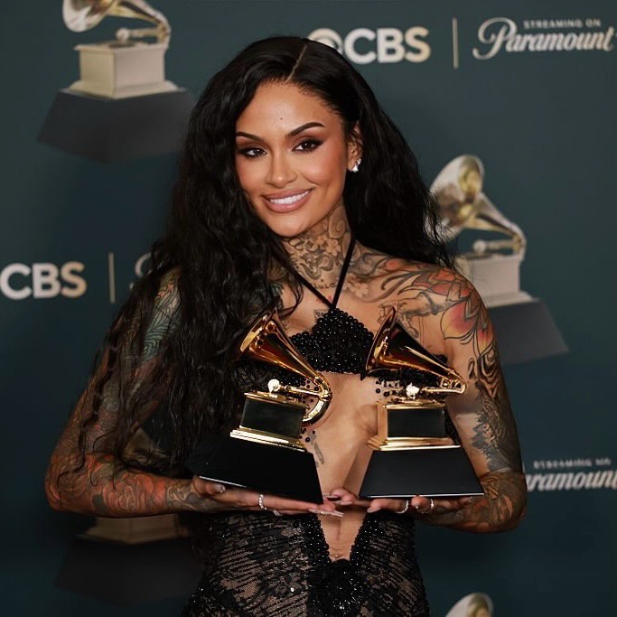 R&B artist Kehlani smiling while holding her two Grammy Awards at the 68th Annual ceremony. She is wearing a sheer black Valdrin Sahiti gown with intricate beading and a central cutout that showcases her tattoos.