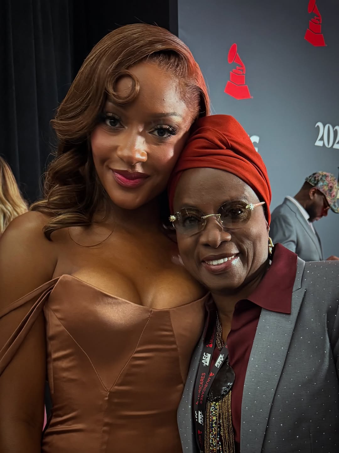 Ayra Starr in a bronze off-the-shoulder gown posing with five-time Grammy winner Angélique Kidjo, who is wearing a grey blazer and red headwrap, at the 2026 MusiCares Person of the Year gala in Los Angeles.