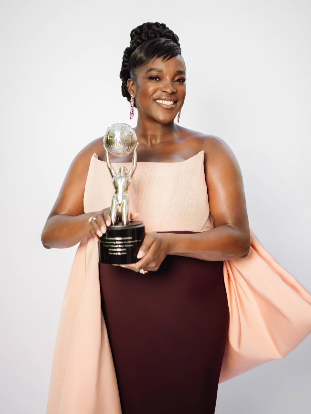 Wunmi Mosaku holding her trophy for Best Supporting Actress at the 57th NAACP Image Awards while wearing a custom blush and burgundy Christian Siriano gown