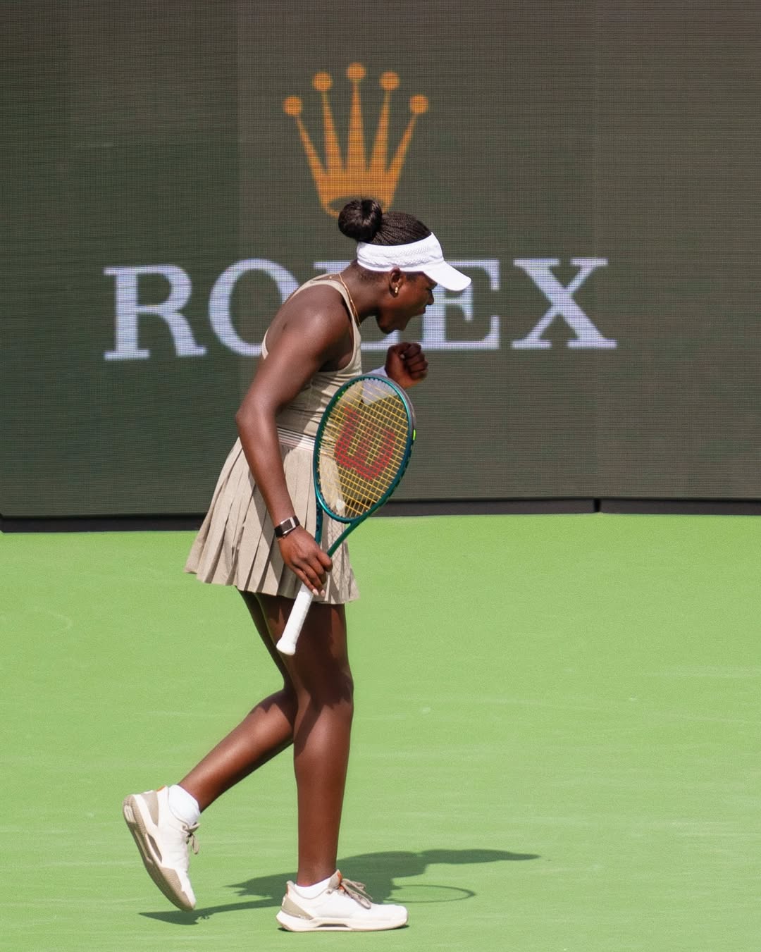 Victoria Mboko in a tan tennis outfit and white visor, pumping her fist in celebration during her match against Amanda Anisimova at the Indian Wells 2026 tournament.
