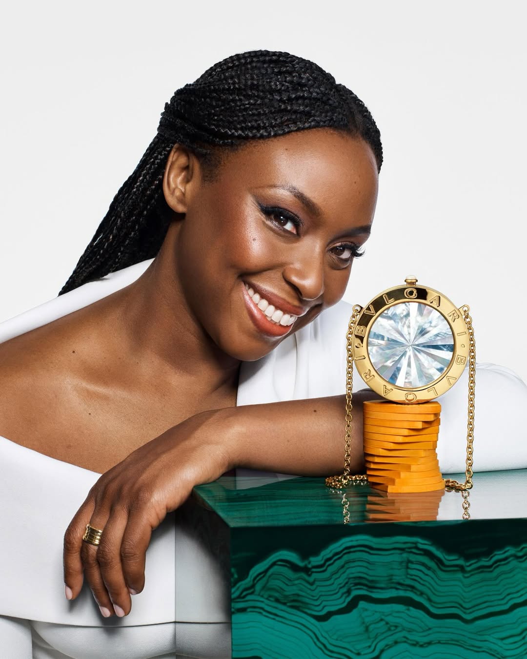 Chimamanda Ngozi Adichie wearing a structured white off-the-shoulder top and gold B.zero1 rings, posed with the circular gold BVLGARI BVLGARI Icons Minaudière and a stack of miniature books.