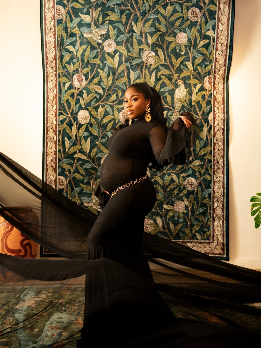 Fashion designer Veekee James posing in a black sheer mesh gown that highlights her baby bump, styled with a gold charm belt and ornate earrings.