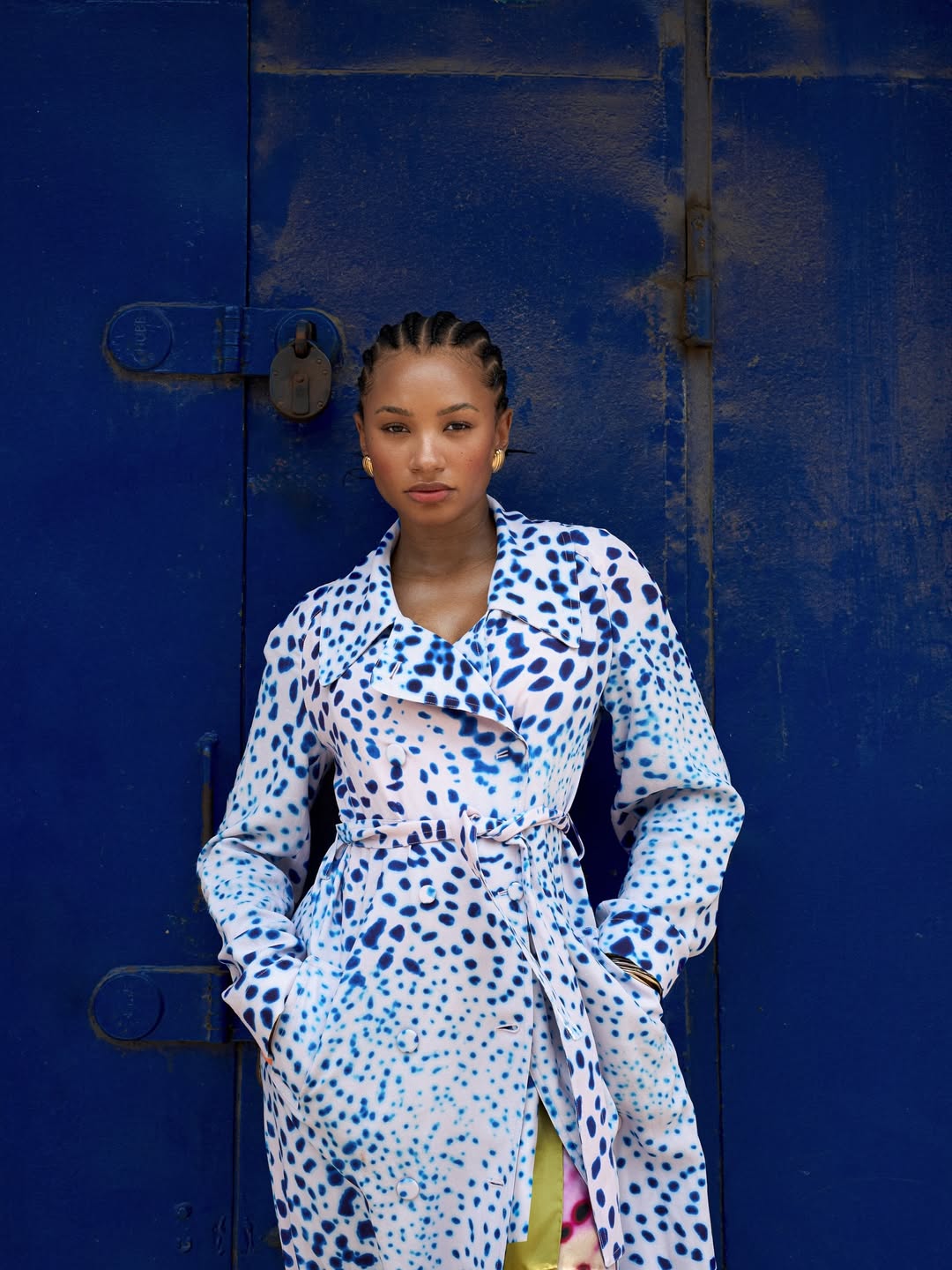 Temi Ajibade in Accra wearing a blue and white dalmatian-print maxi coat dress and neat back-braided cornrows, posing against a cobalt blue metal door.