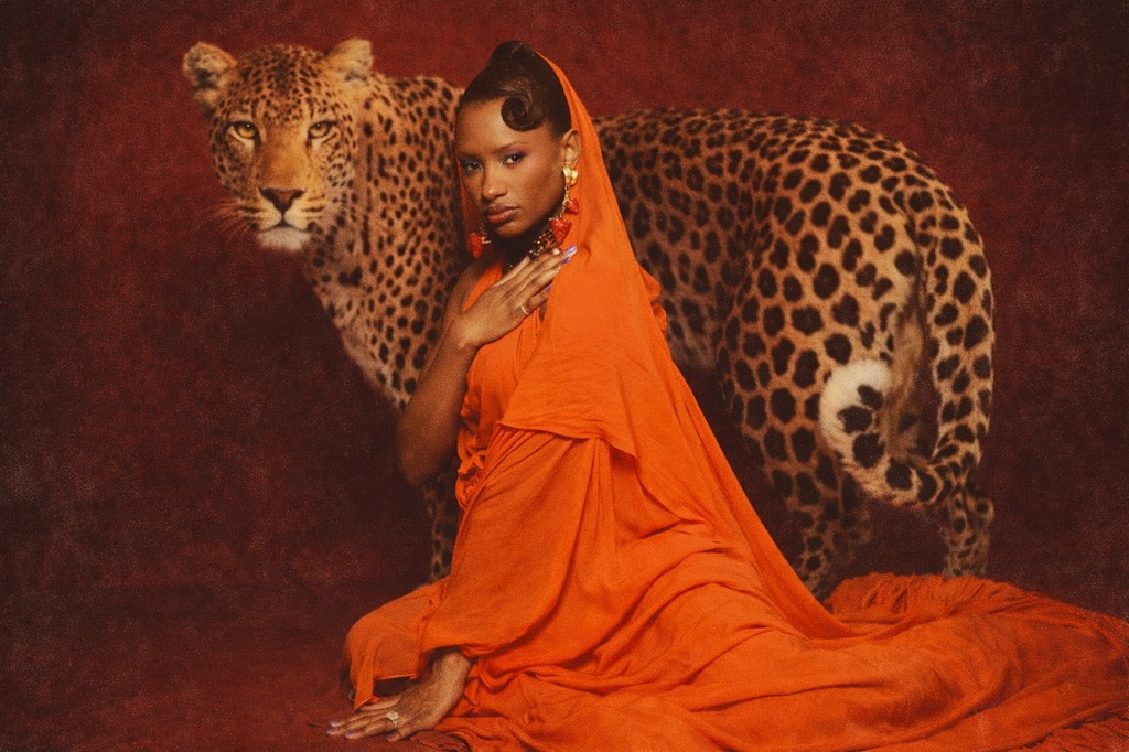 Temi Ajibade (Otedola) wearing a head-to-toe draped orange gown with a matching veil, posing with a leopard against a deep burgundy textured backdrop for her 30th birthday shoot.