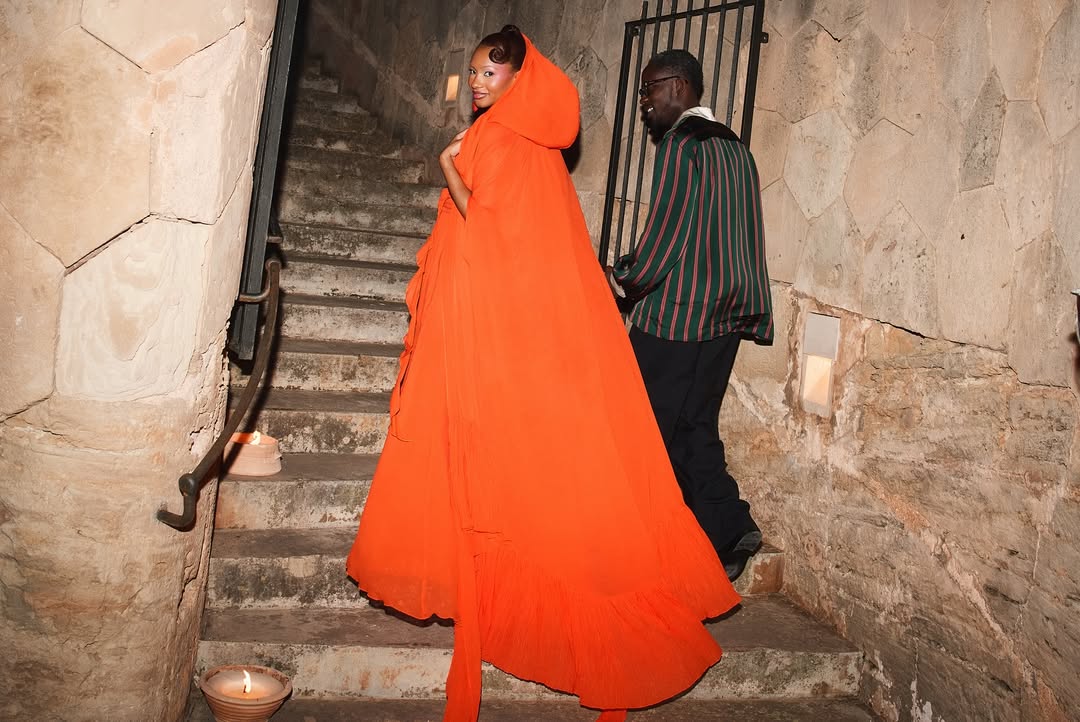 Temi Ajibade in a voluminous draped orange gown walking up stone stairs alongside her husband, Tosin Ajibade (Mr Eazi), who is wearing a green and red striped jacket. Small candles light the stone steps in an intimate, rustic setting.