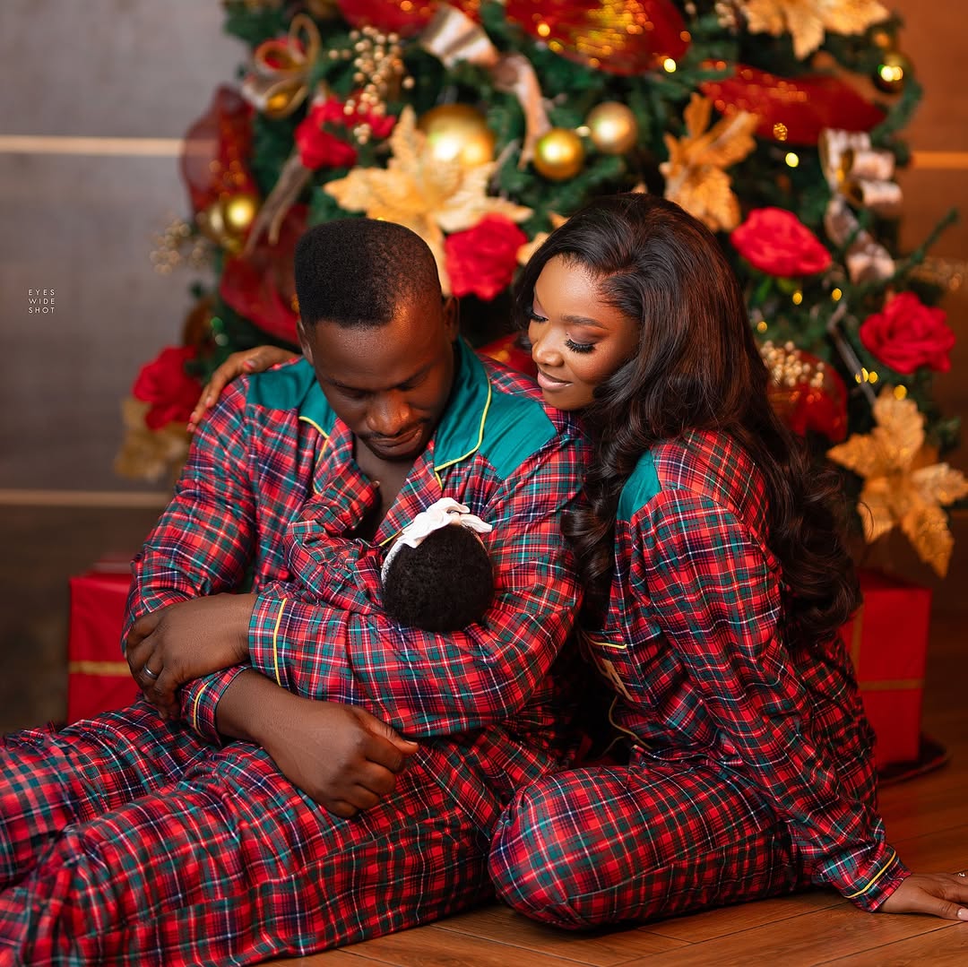 Wofai Fada and Taiwo Cole sitting in front of a decorated Christmas tree in matching red and green plaid pajamas. Taiwo is cradling their newborn daughter, Ifedayo Konaghai Cole, who is wearing a white headband.