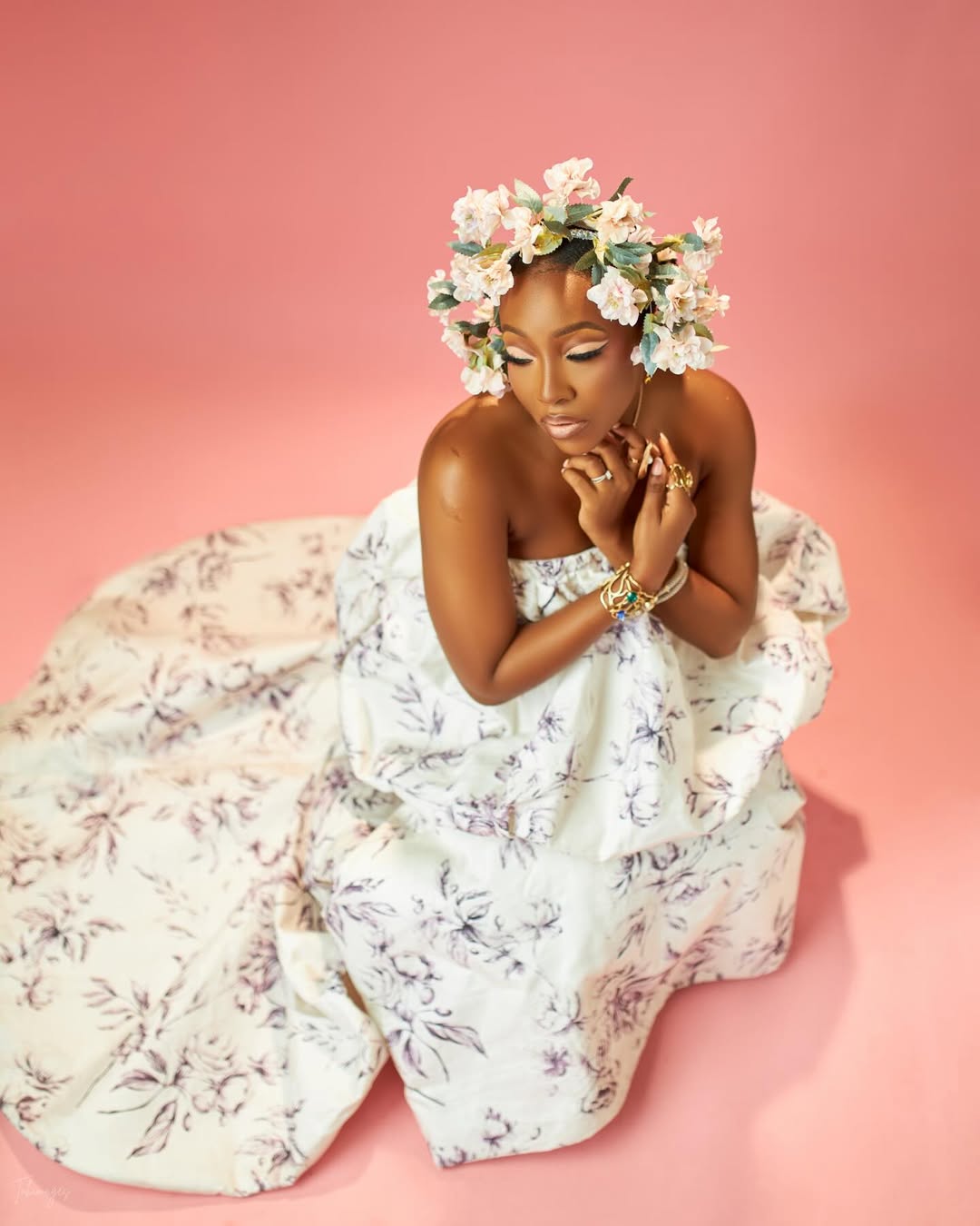 Actor Ini Dima-Okojie sitting in a voluminous white and purple floral strapless gown with a matching floral headpiece against a pink backdrop.