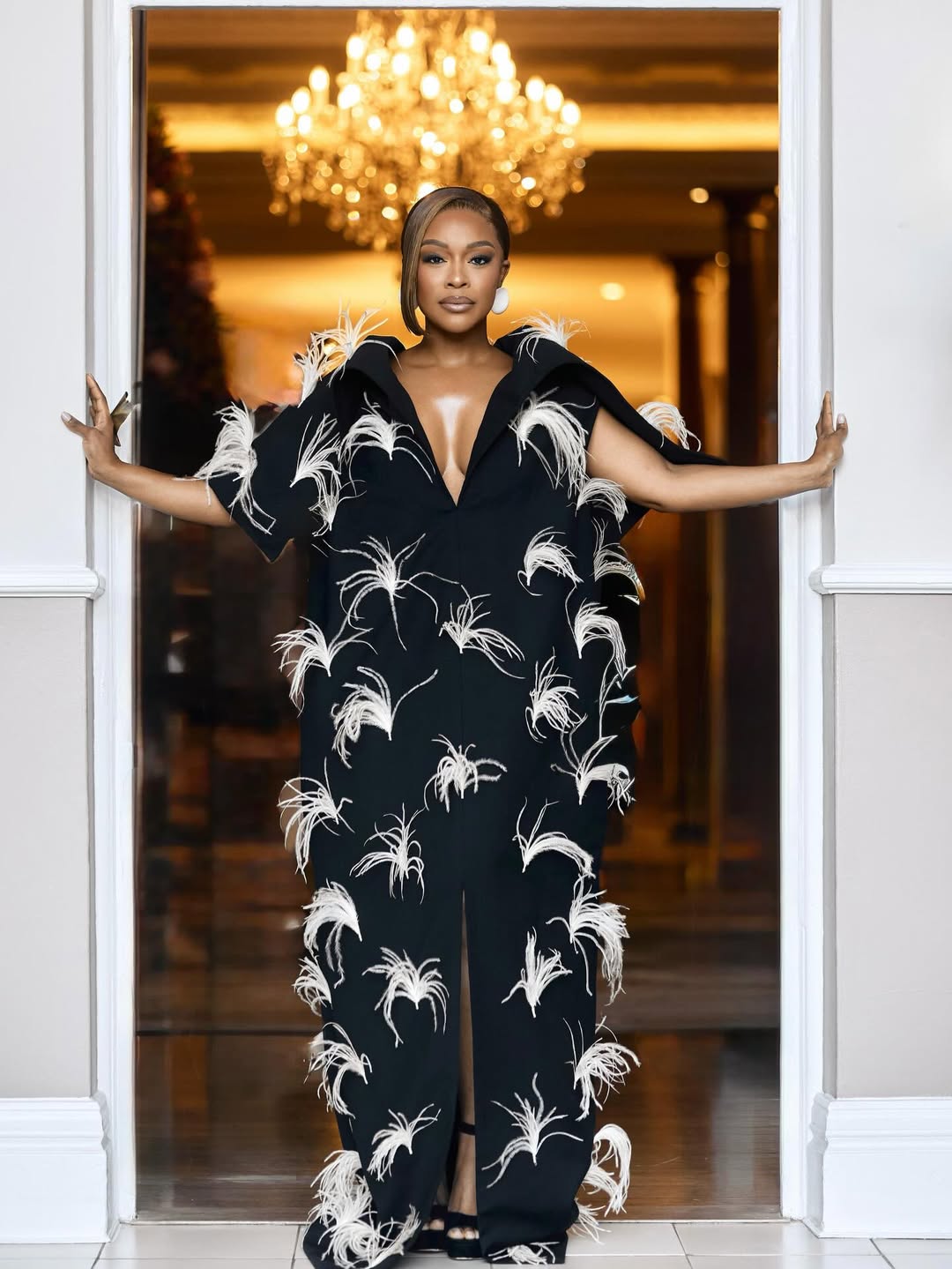 South African actress and AMVCA 12 co-host Nomzamo Mbatha posing in a black floor-length gown adorned with white feathers under a chandelier.
