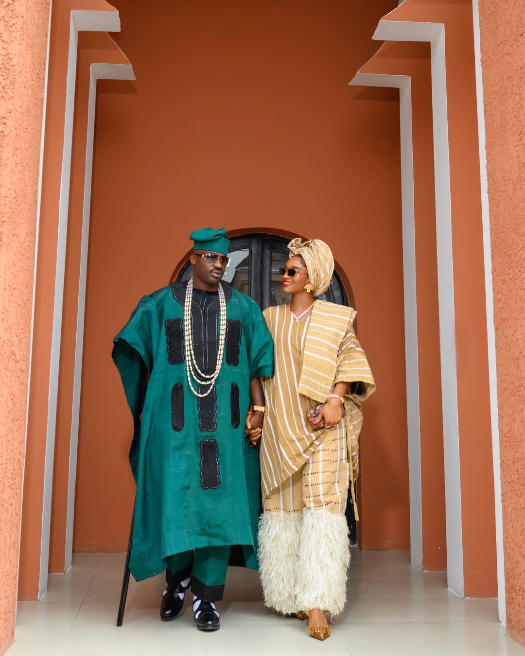 Wofai Fada and husband Taiwo Cole in traditional Nigerian attire. Wofai wears a gold-striped Aso-Oke with feathered fringe sleeves and a matching gele, while Taiwo wears a deep green Agbada with black embroidery, beads, and a matching fila.