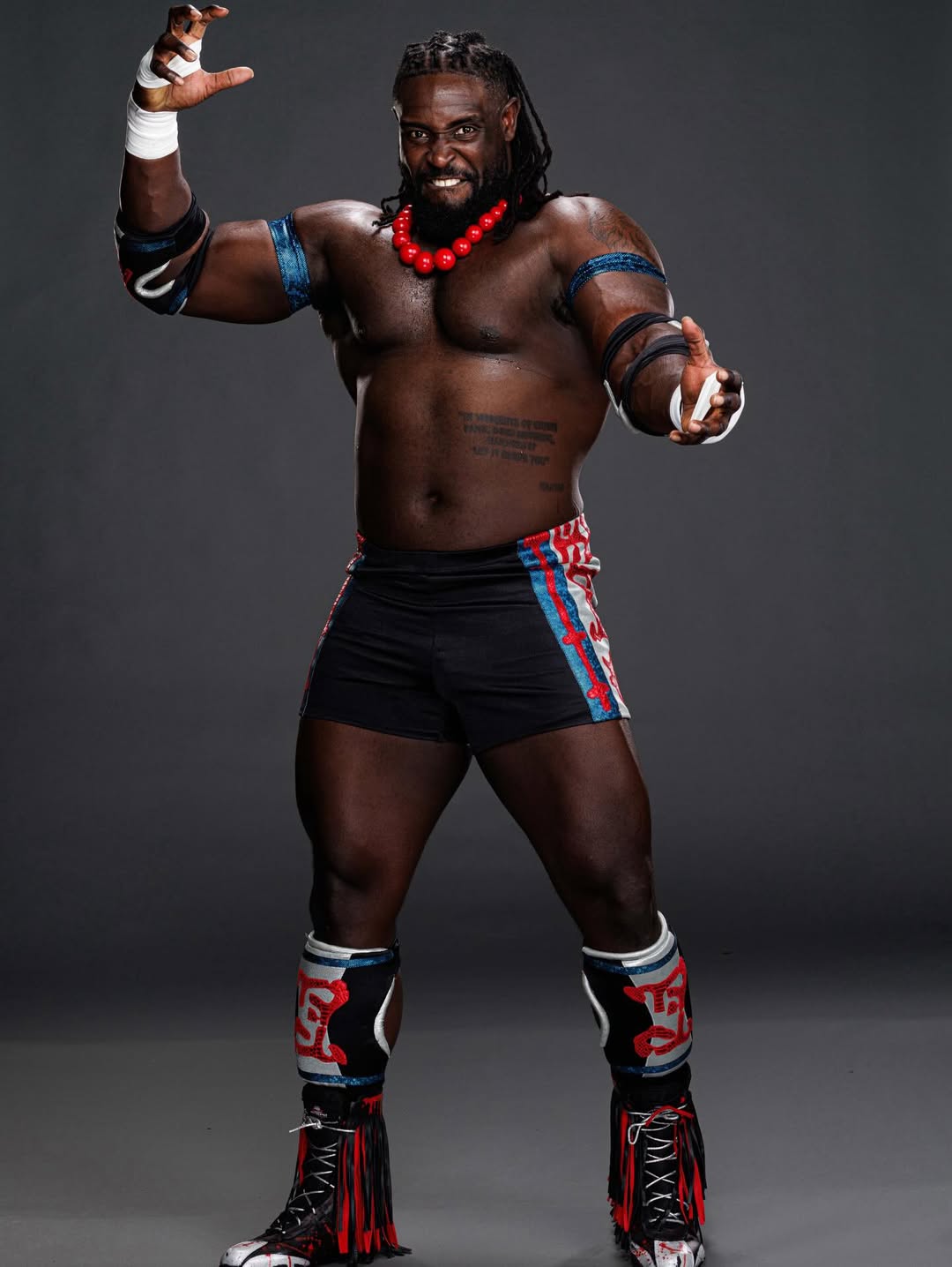 A studio portrait of Nigerian WWE wrestler Oba Femi, "The Ruler," standing in his signature wrestling gear with a red beaded necklace and blue armbands.