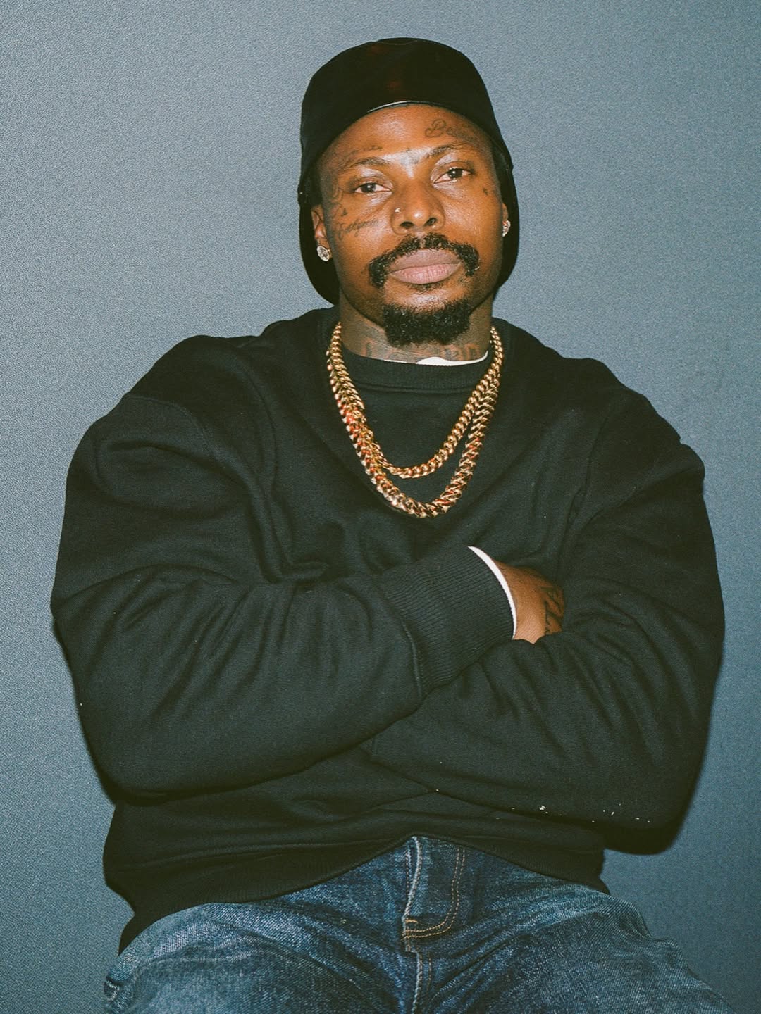 A studio portrait of Asake wearing a black sweatshirt, two gold Cuban link chains, and a black beanie, posing with his arms crossed against a neutral grey background.