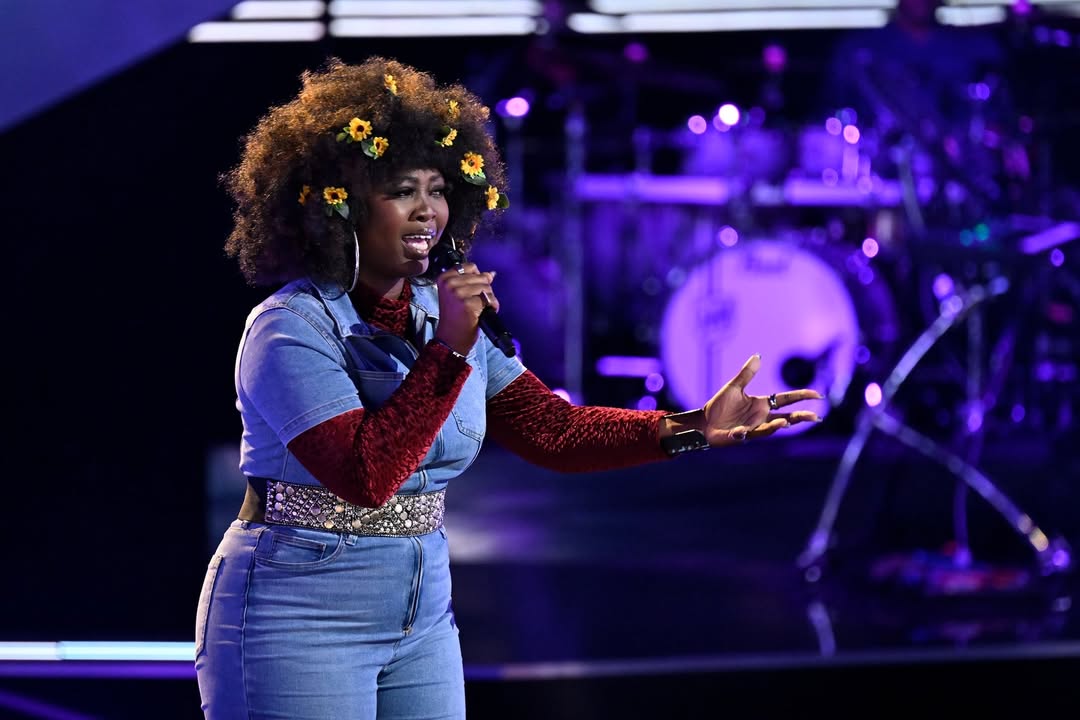 Alexia Jayy performing on The Voice stage in a denim jumpsuit and red textured sleeves with sunflowers in her hair.
