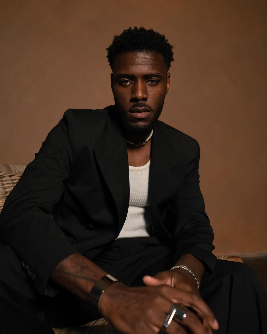 Actor Tega Alexander, who joins Bridgerton Season 5 as Christopher Anderson, posing in a black tailored blazer and pearl necklace.
