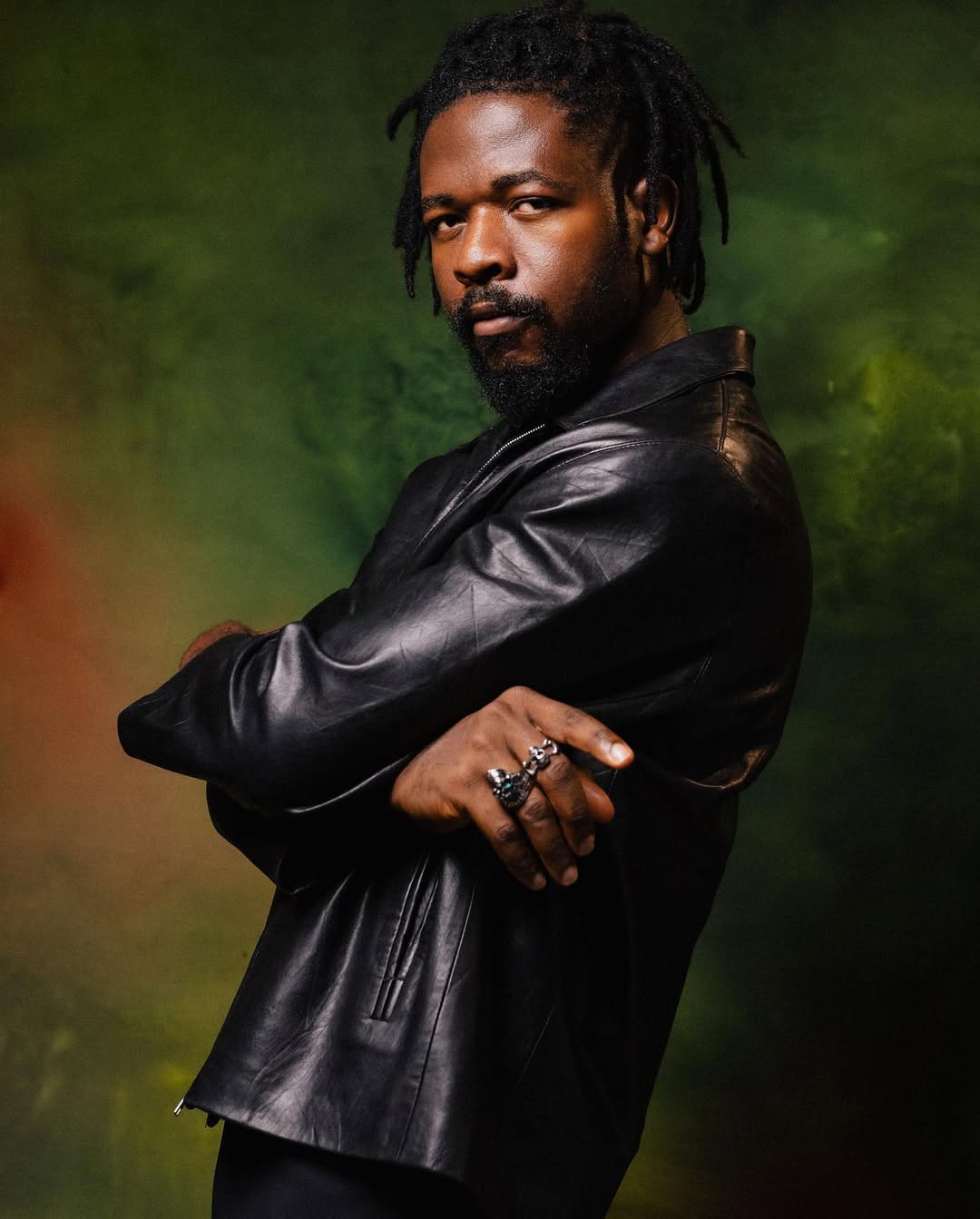 Nigerian singer-songwriter Johnny Drille posing in a black leather jacket with his arms crossed against a green textured background.
