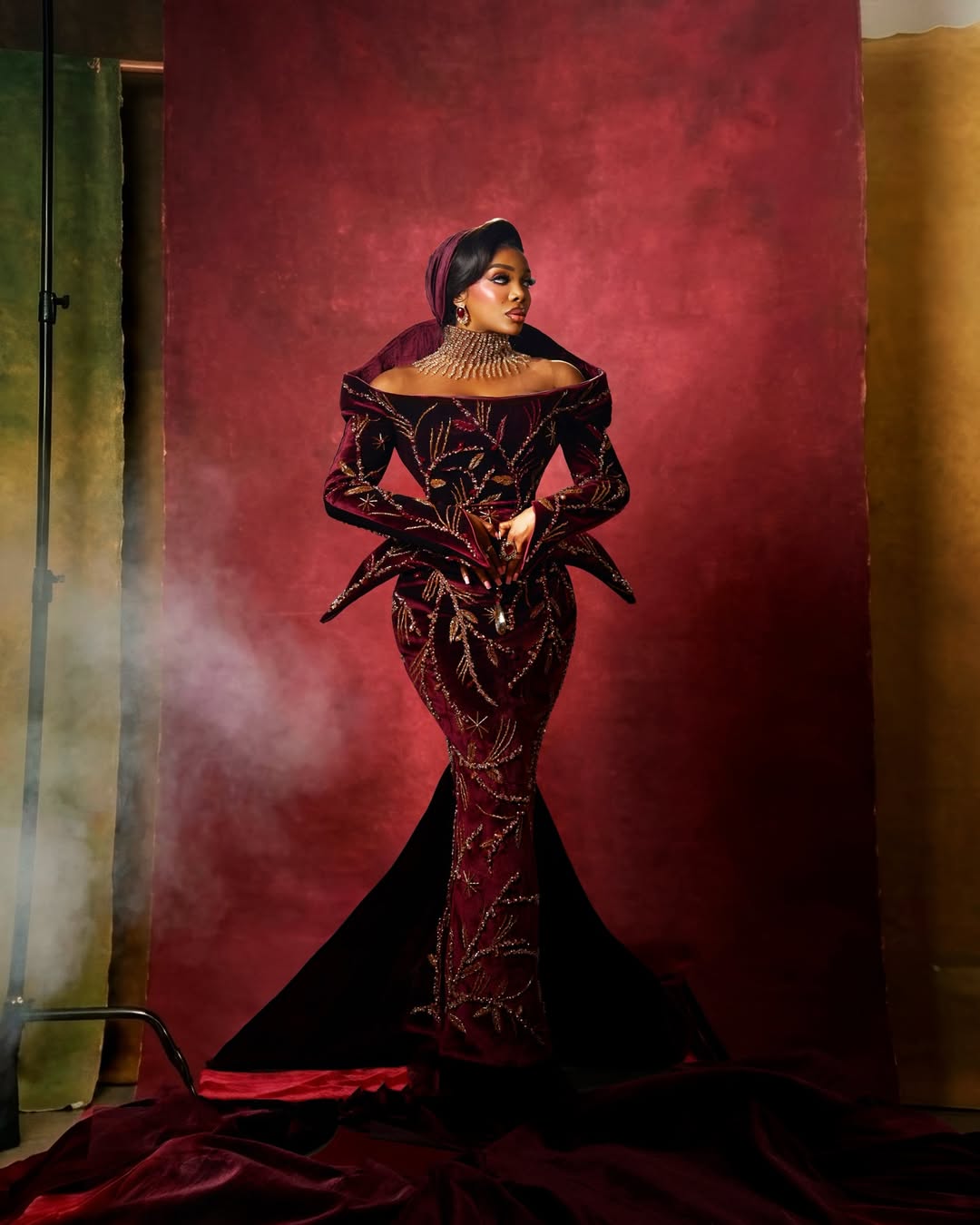 Chioma Ikokwu in a deep burgundy velvet mermaid gown by Sevon Dejana, featuring off-shoulder sculptural wings at the shoulders and intricate gold embroidery. Photographed by Ayo Makinwa against a smoky red backdrop.