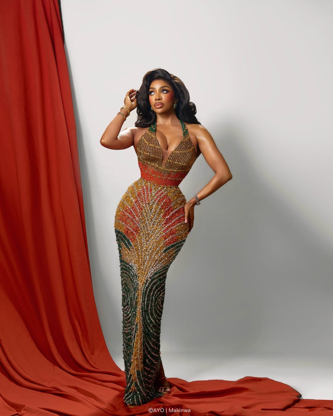 Chioma Ikokwu poses in a fitted, floor-length beaded halterneck gown by Djec Fashion, featuring concentric swirls of gold, red, orange, and green that move around the body like a map.