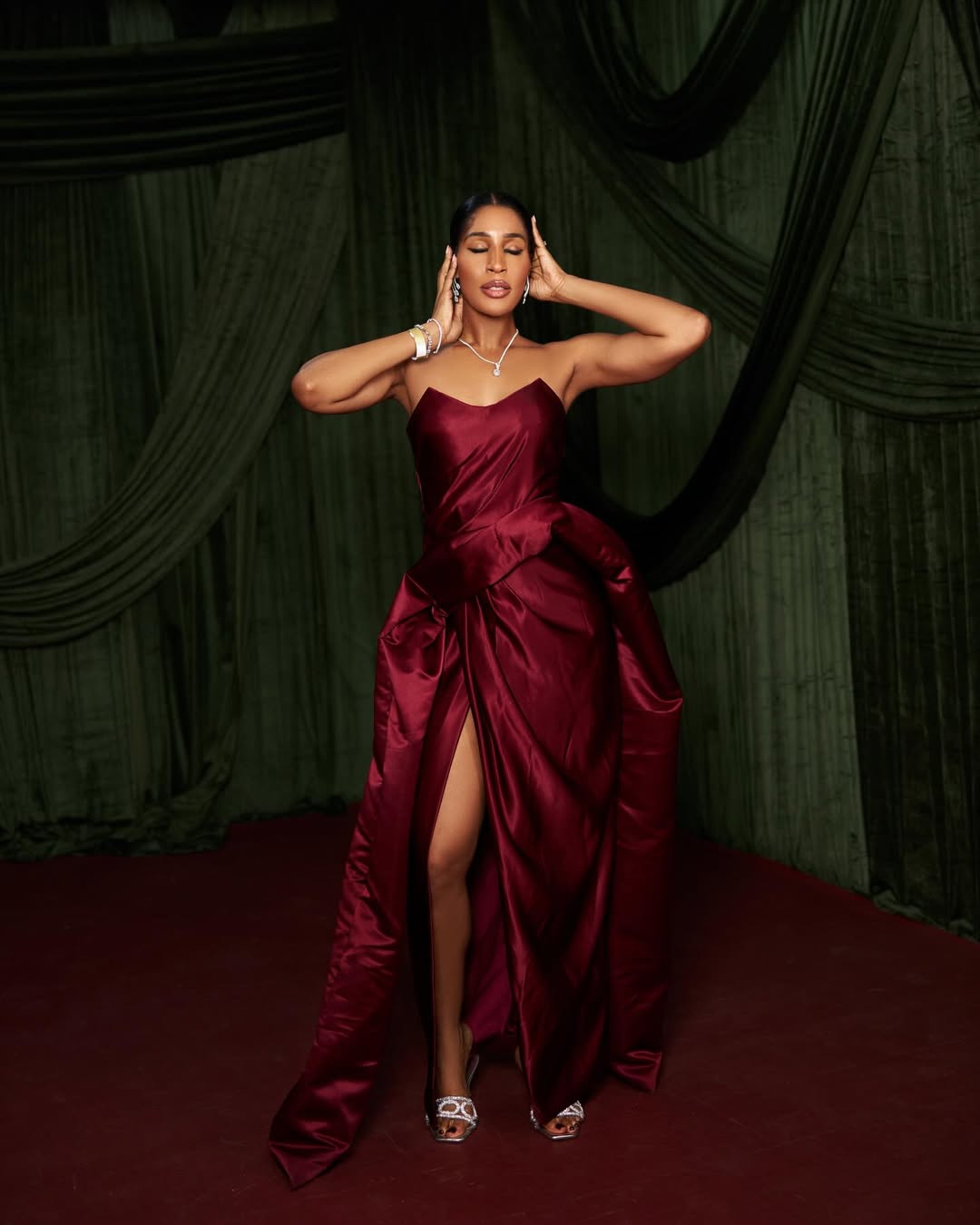 Nicole Chikwe posing in a strapless burgundy satin gown with a thigh slit and sweeping train against a dark green velvet backdrop.