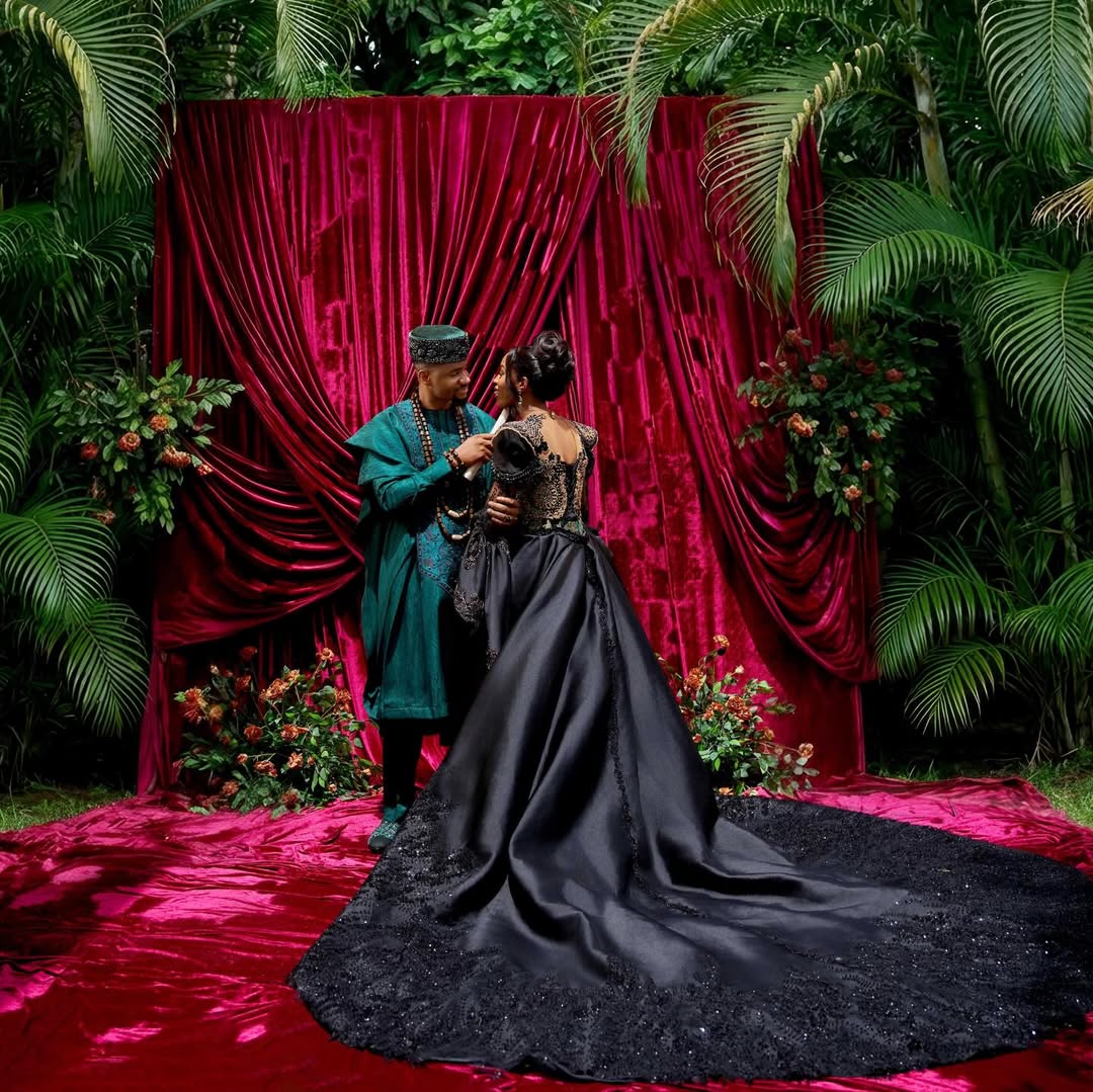 Jide Kene Achufusi and Ifeoma posing against a draped magenta velvet backdrop at their traditional wedding, showcasing Ifeoma's dramatic circular black satin train and Isi-Agu detailing.