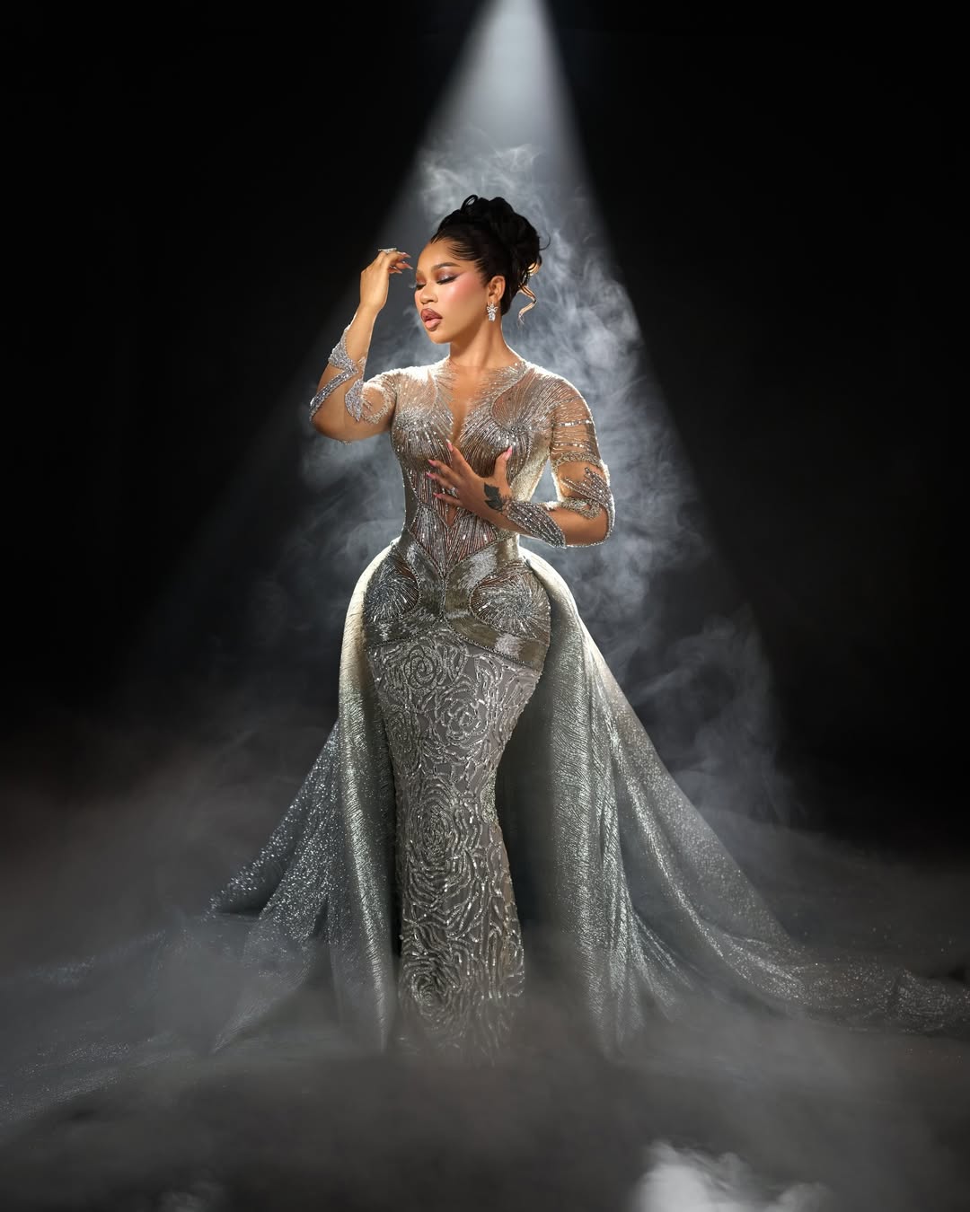 Diiadem in a silver and nude illusion mermaid gown with long sheer sleeves, dense beadwork, and a sweeping overskirt against a dark, smoky background.