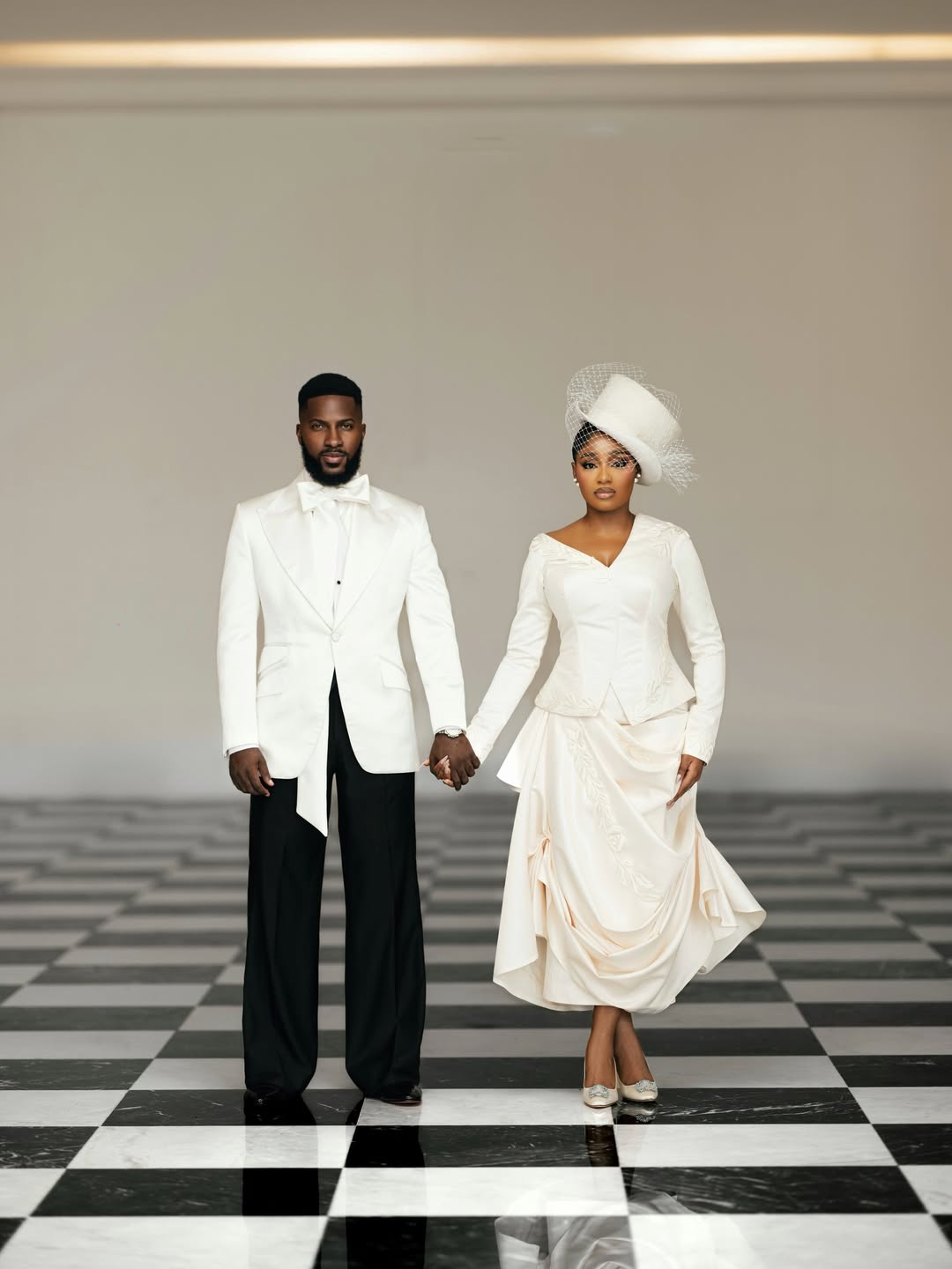 A formal portrait of Akin Faminu in a custom Deji and Kola tuxedo and Kiky Festus in a Mazelle Bridal two-piece set, holding hands inside a grand hall with checkered marble flooring and tall white columns.