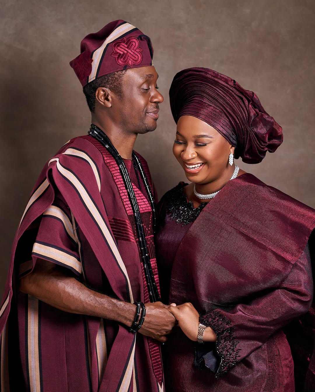 Nathaniel Bassey and his wife Sarah in a pose, featuring his burgundy Agbada with tone-on-tone embroidery and her matching embellished Iro and Buba with a crown-style Gele.