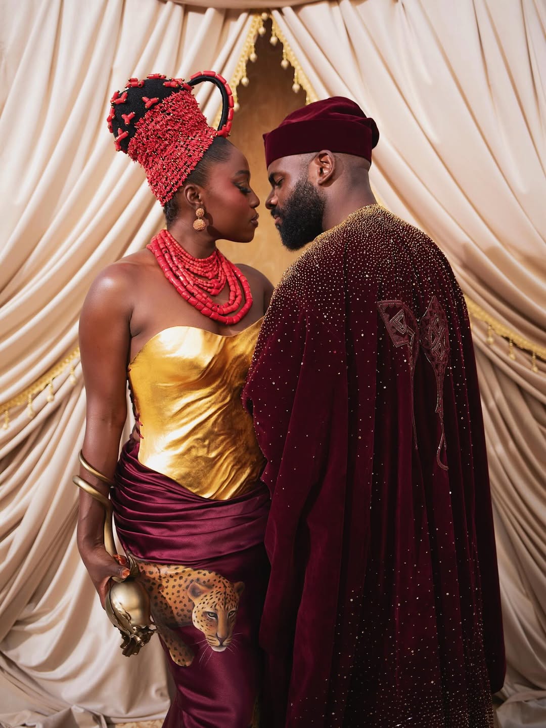 Fisayo Longe wearing a gold metallic breastplate corset and maroon leopard-print skirt alongside Afolabi Mosuro in a velvet maroon Agbada.