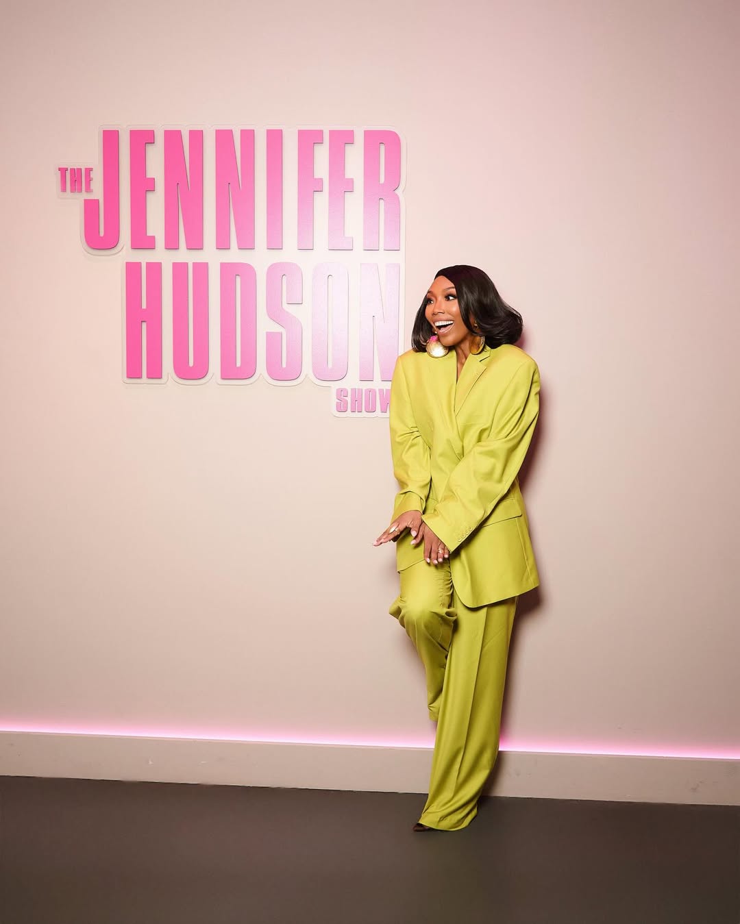 Brandy posing in a full-length monochrome lime green power suit consisting of an oversized blazer and wide-leg trousers against a pink JHud Show logo wall.