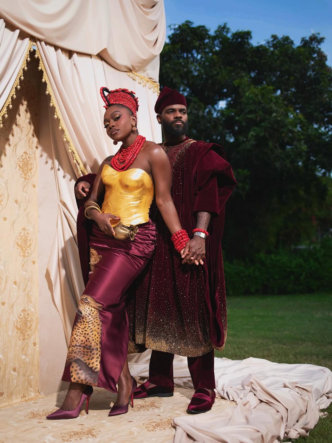Fisayo Longe wearing a gold metallic breastplate corset and maroon leopard-print skirt alongside Afolabi Mosuro in a velvet maroon Agbada.