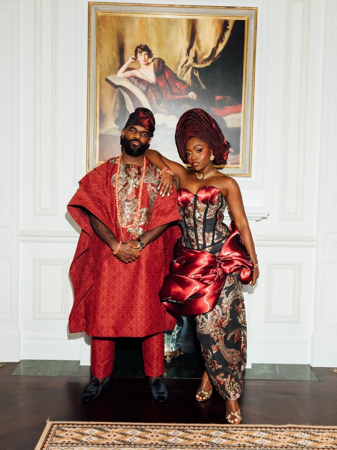 Fisayo Longe in a strapless crimson and black brocade gown with a structural bow, standing with Afolabi Mosuro in a matching Agbada in front of a white wall.