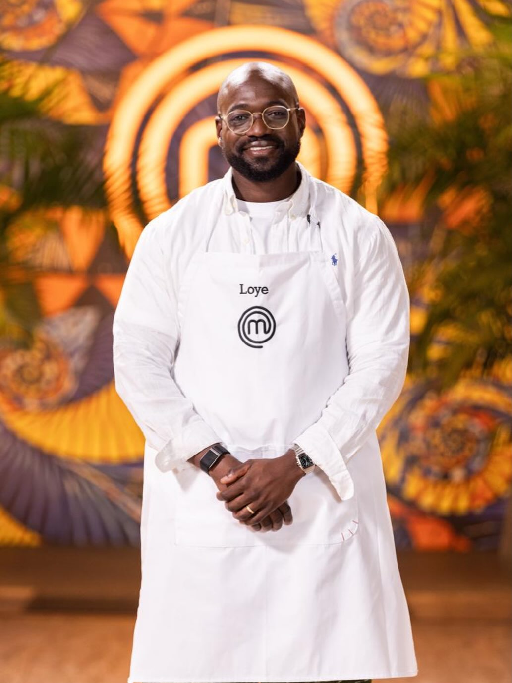 MasterChef Nigeria contestant Loye Oyedotun smiling with hands clasped in front, wearing a white apron, glasses, and a white button-down shirt.