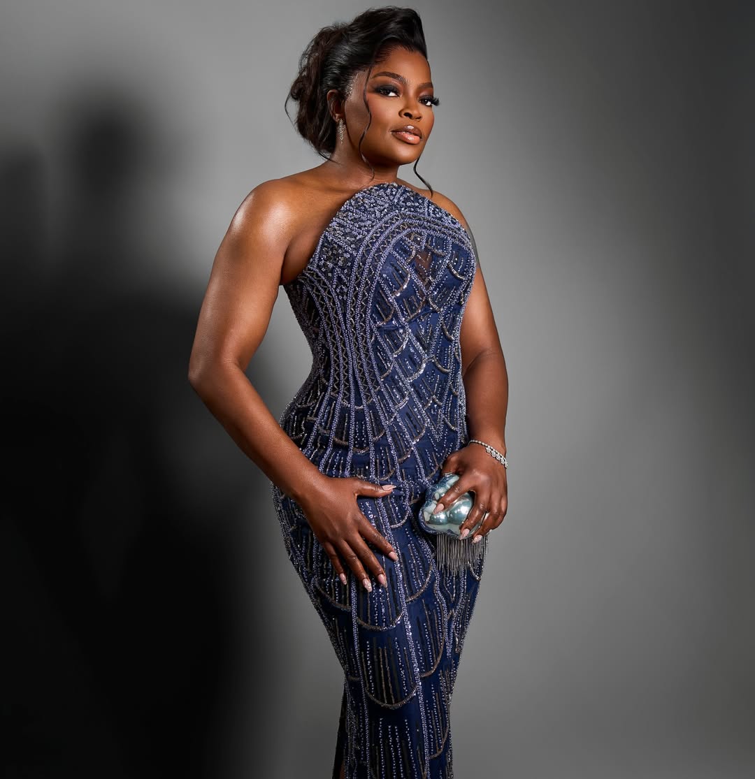 A portrait of Funke Akindele showing a sleek high updo, neutral makeup, and a small metallic silver clutch held against the beaded navy dress.