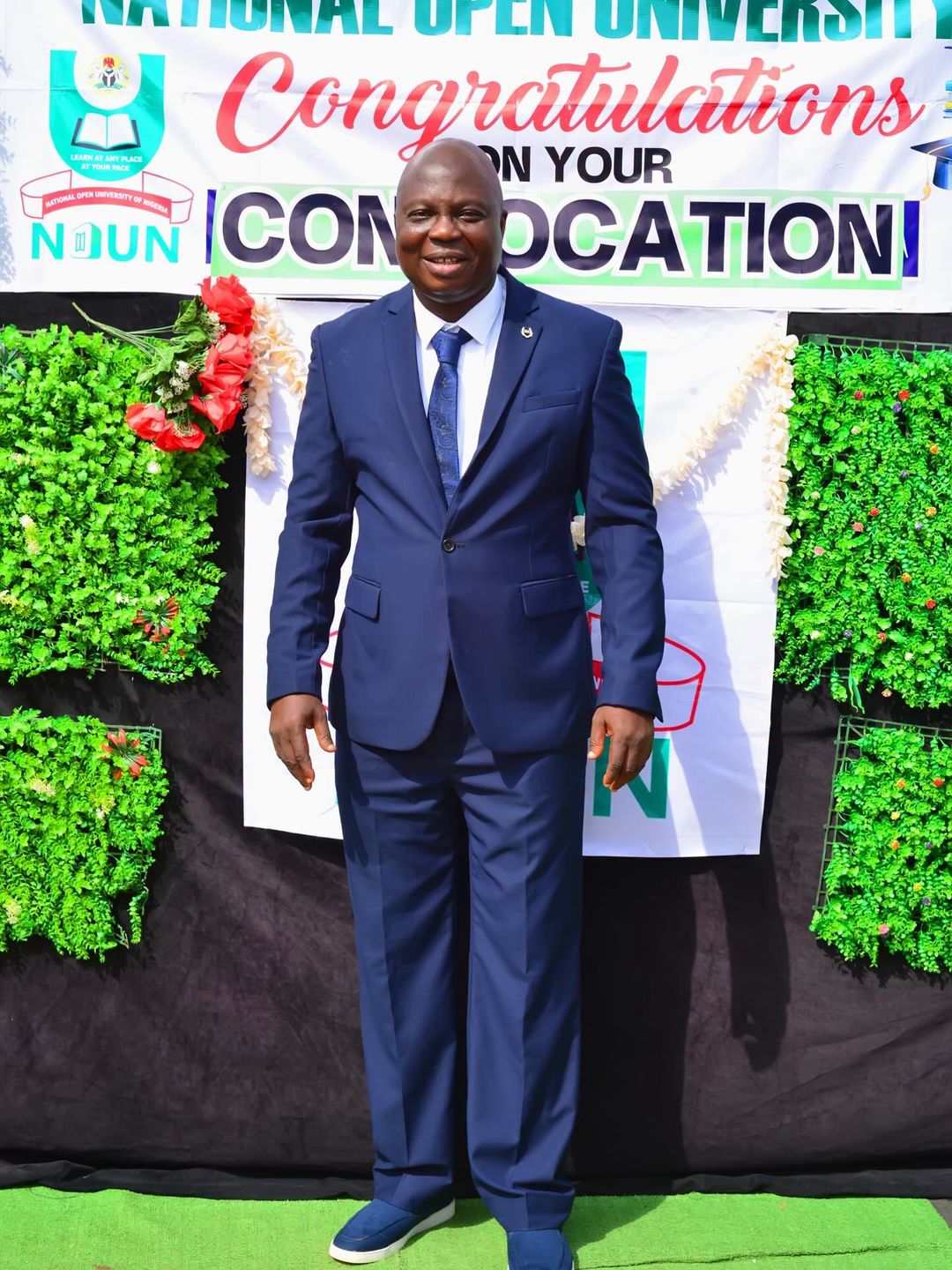 Nollywood actor Bolaji Amusan (Mr Latin) in a navy blue suit standing in front of a National Open University of Nigeria (NOUN) graduation banner.