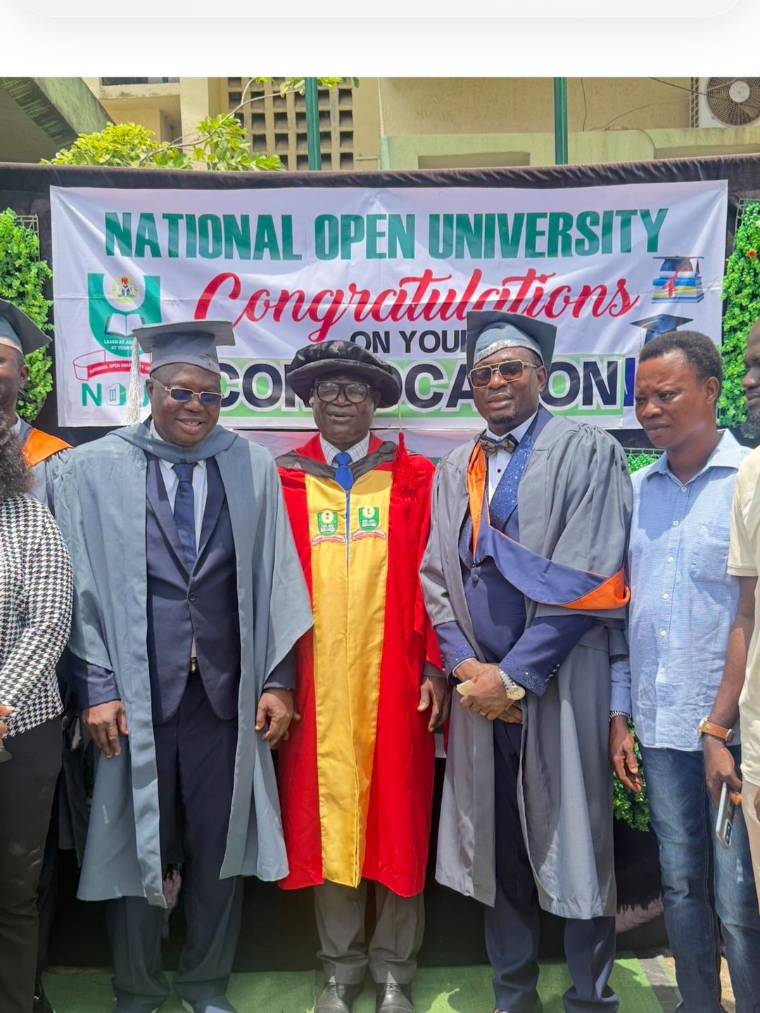 Nollywood veteran Bolaji Amusan (Mr Latin), in a blue graduation gown and cap, stands smiling while holding his certificate with a fellow female NOUN graduate at the 15th Convocation Ceremony.