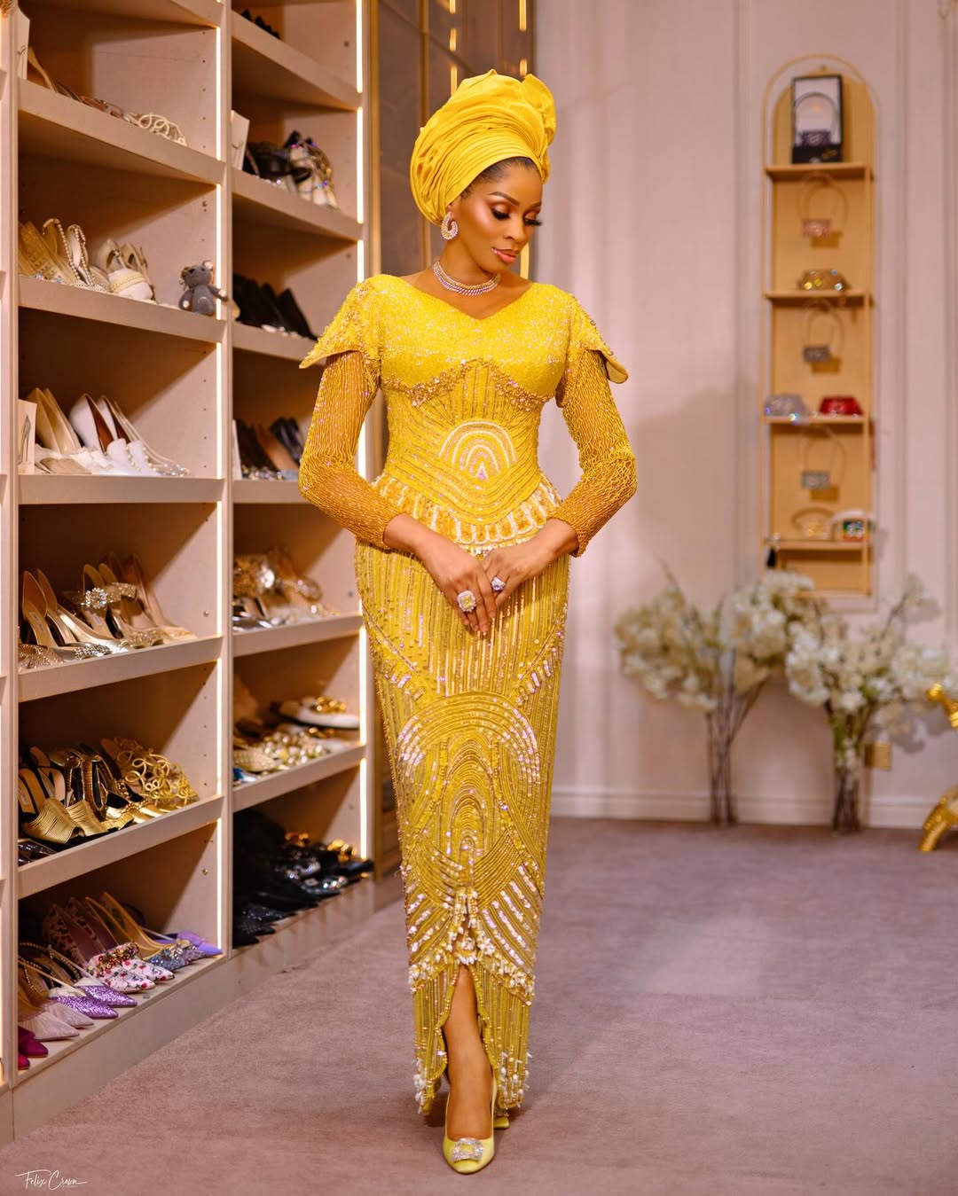 Shade Okoya in a luxury walk-in closet, wearing a beaded yellow lace dress with sheer sleeves and a fringe peplum detail.