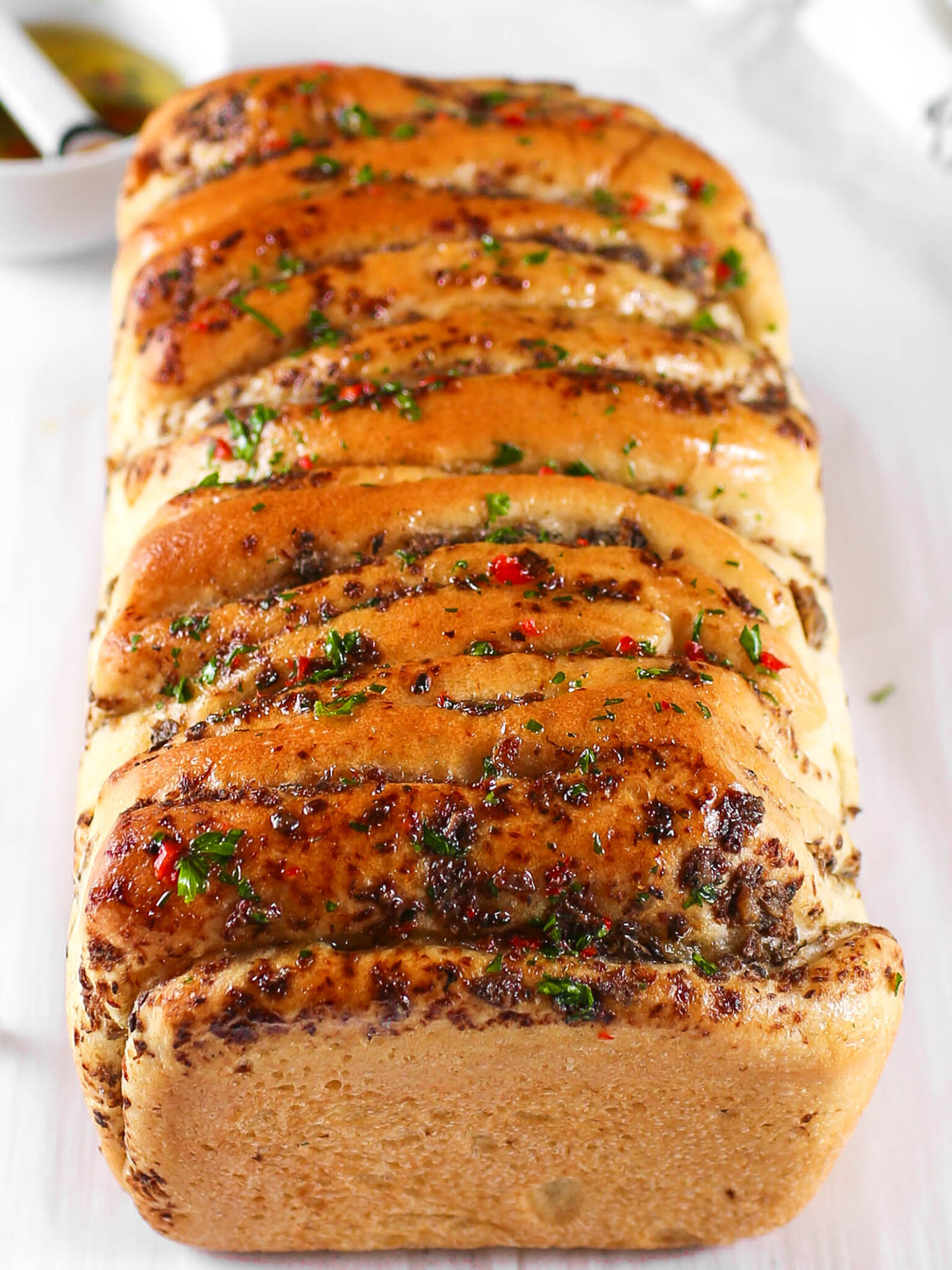 A golden-brown, freshly baked loaf of Kikifoodies' sardine pull-apart bread brushed with glossy, spicy oil and topped with chopped parsley and red pepper flakes.
