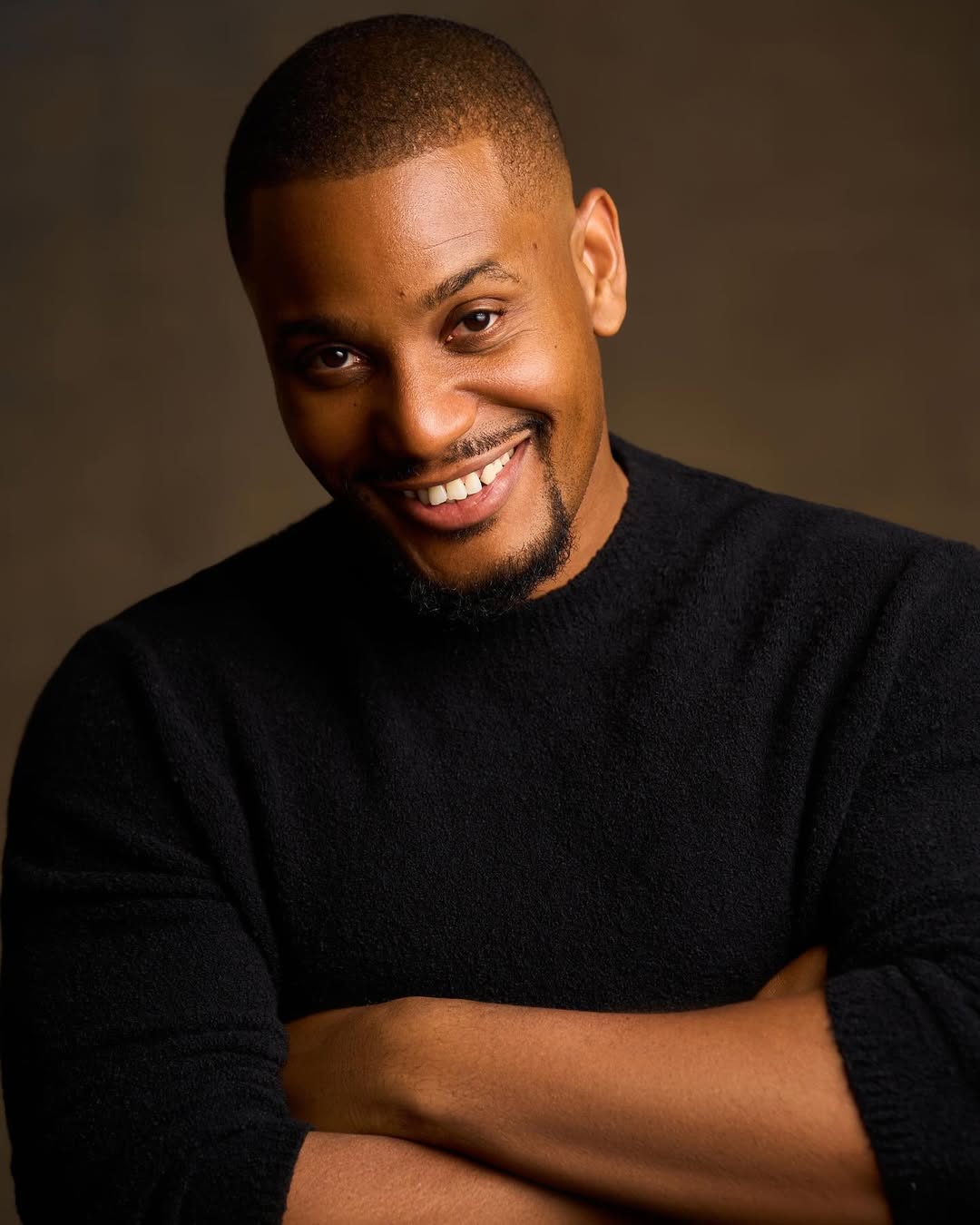 A close-up studio portrait of Nollywood actor Alexx Ekubo smiling and wearing a black jumper.