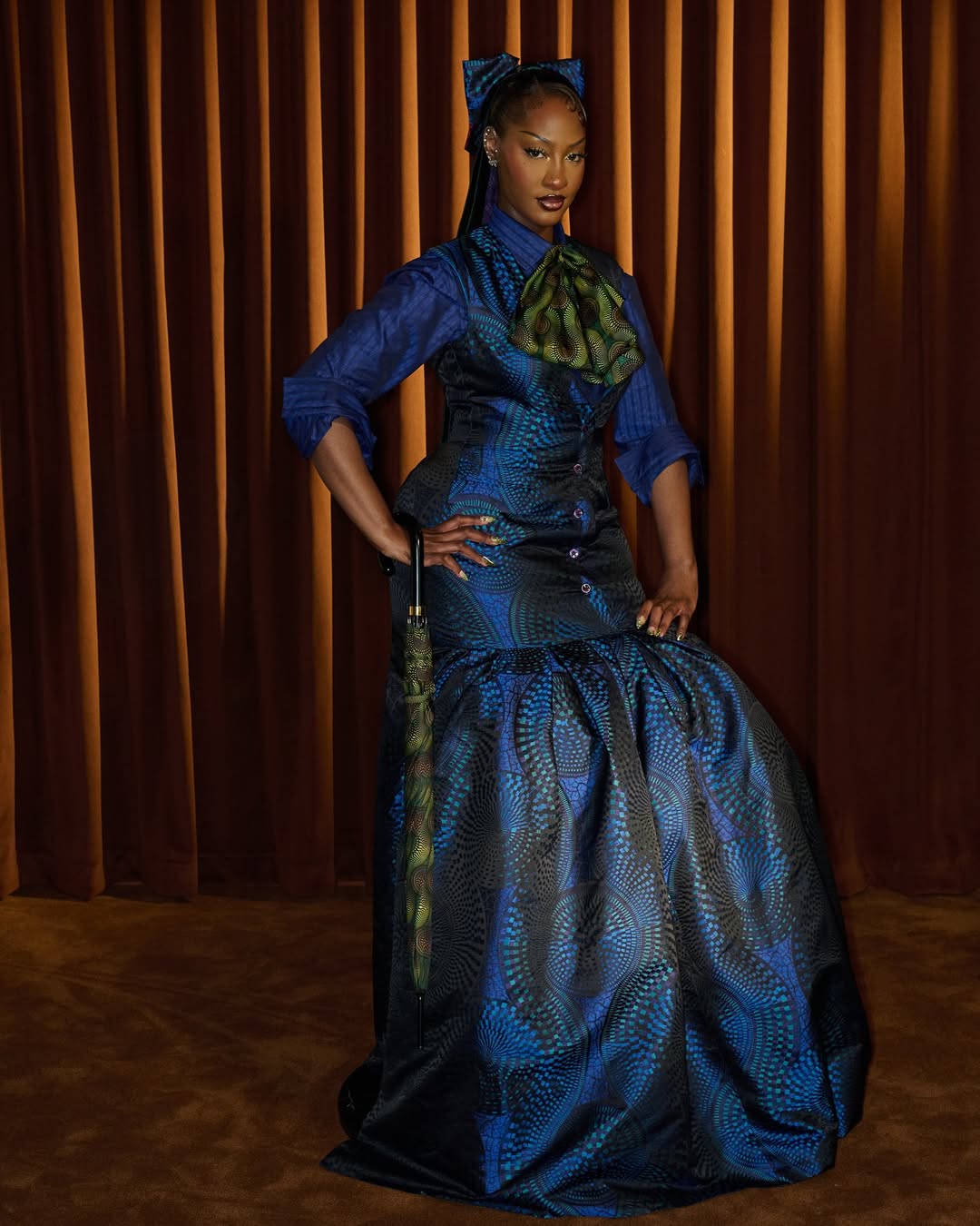 Tems in a blue patterned Ozwald Boateng dress with an umbrella accessory for the 2025 Met Gala "Superfine" theme.