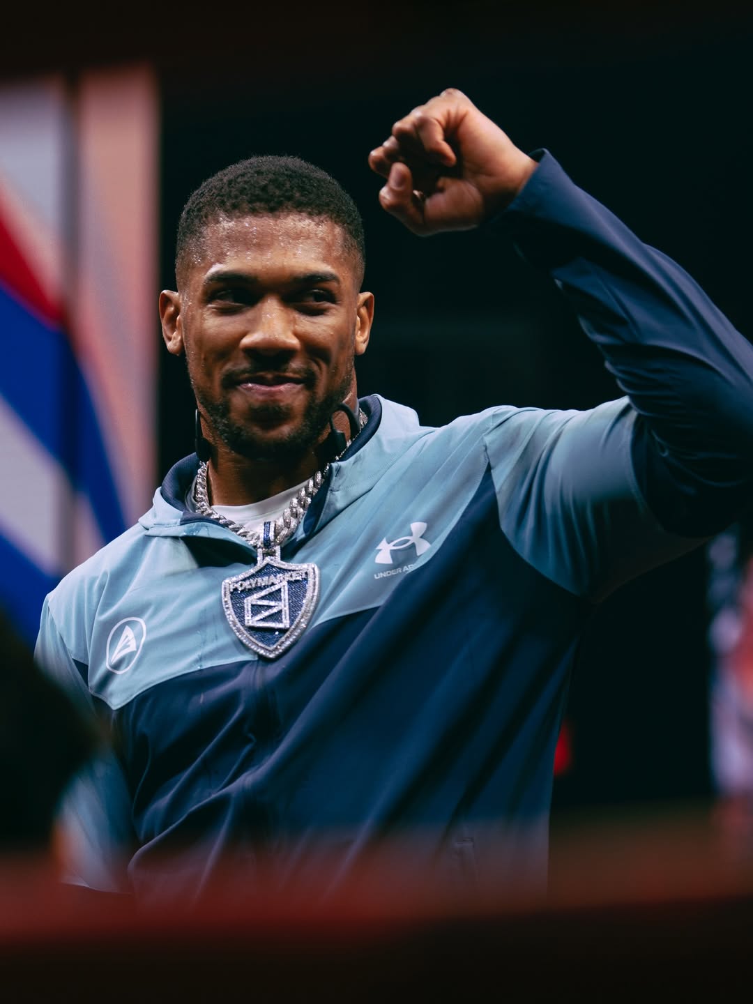 Anthony Joshua smiling and raising a fist while wearing an Under Armour tracksuit and a large Polymarket diamond chain.