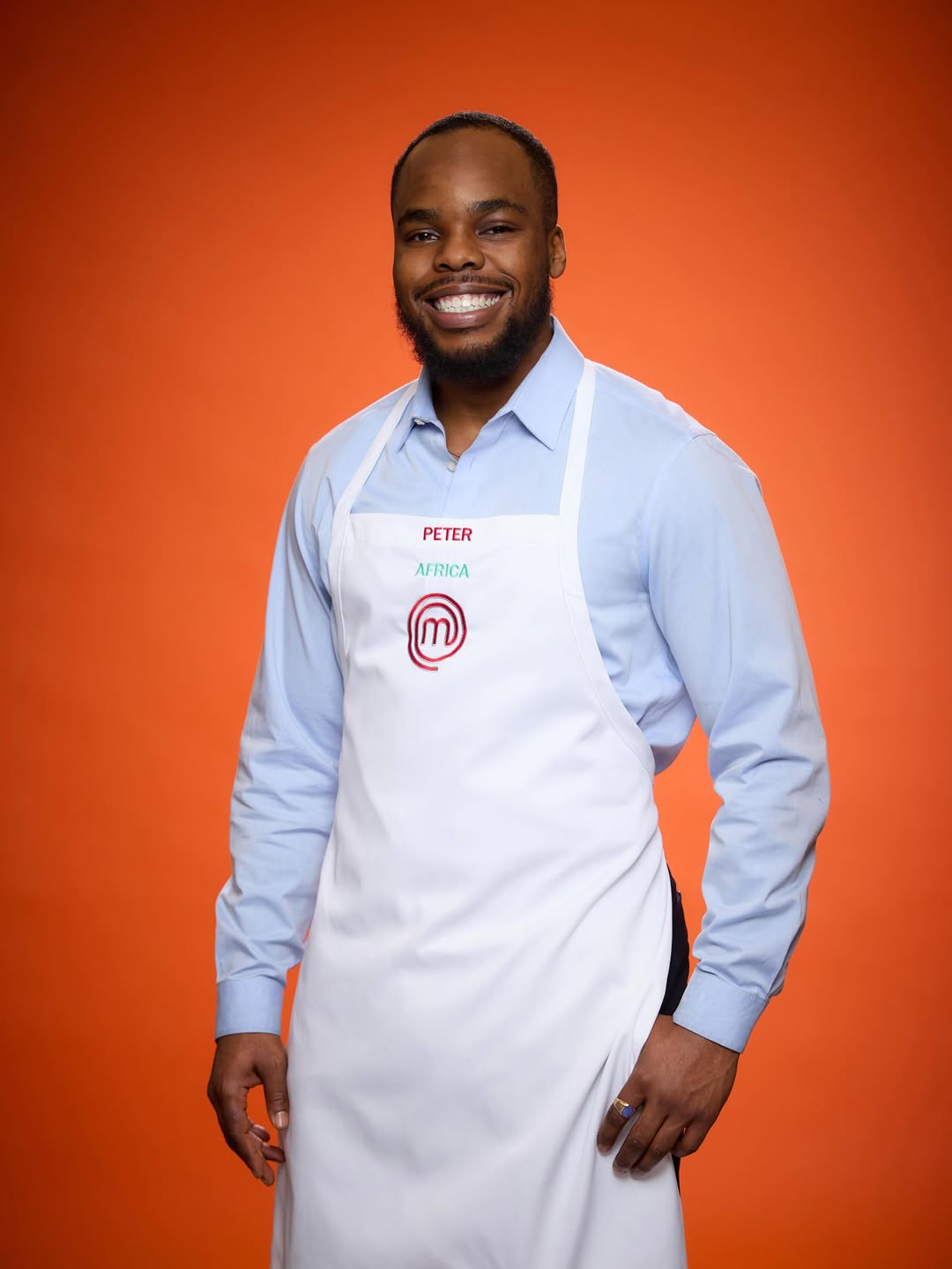 Peter Egede, a Nigerian contestant on MasterChef Season 16: Global Gauntlet, smiling in a white MasterChef apron with "Africa" embroidered in green.