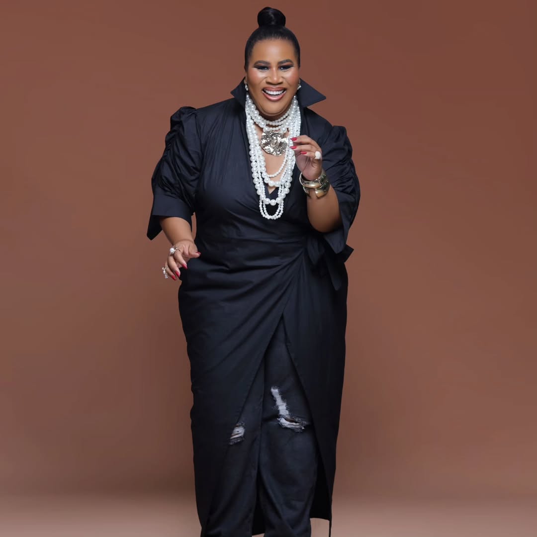 Chioma "Chigurl" Omeruah laughing in a full-length 50th birthday portrait, wearing a sculpted black wrap top with ruched sleeves, distressed black jeans, and layered oversized pearls.