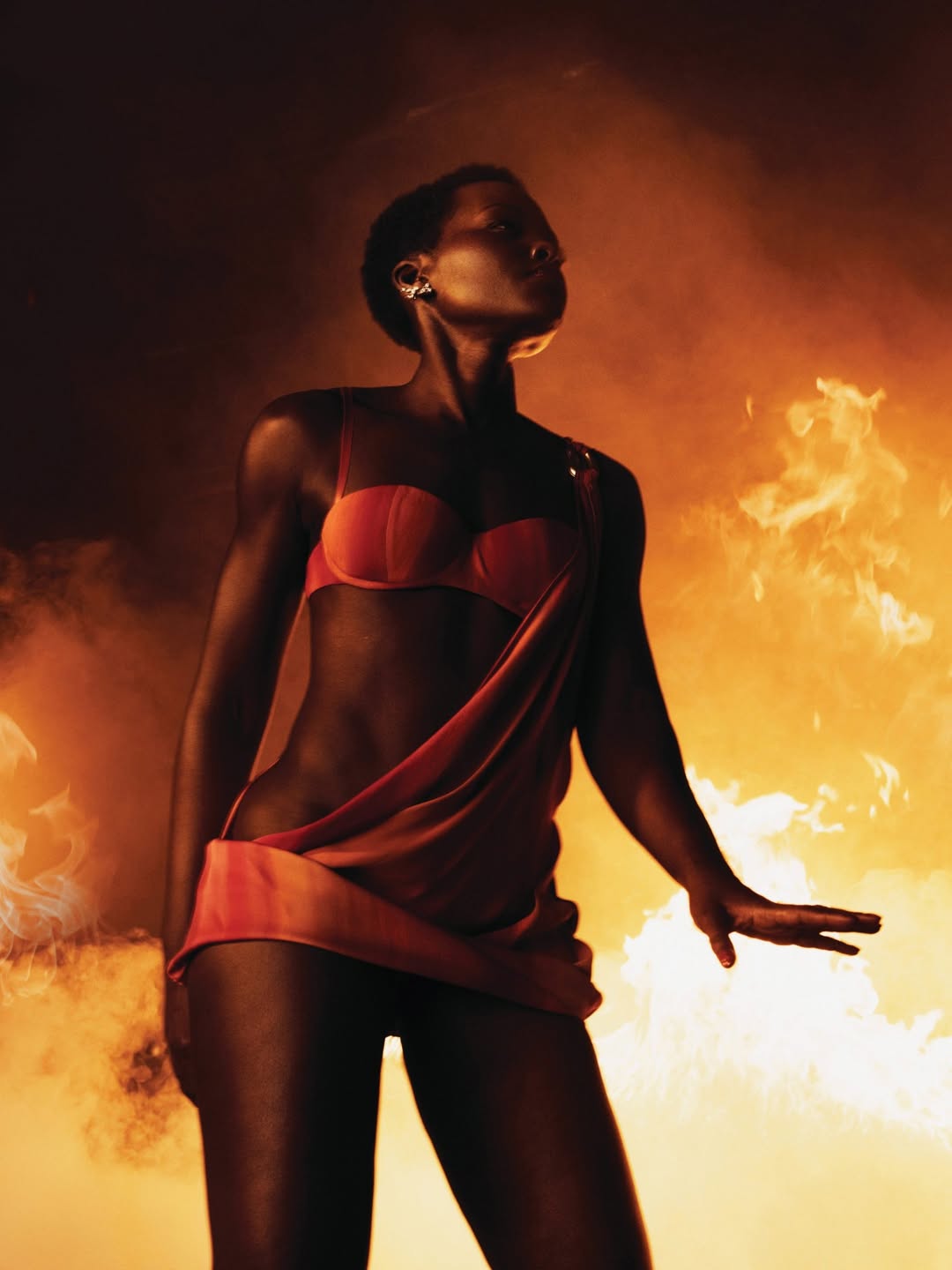 Lupita Nyong'o poses in an asymmetrical orange and red LaQuan Smith dress for ELLE Magazine’s Summer 2026 issue, styled by Law Roach with a background of studio flames.