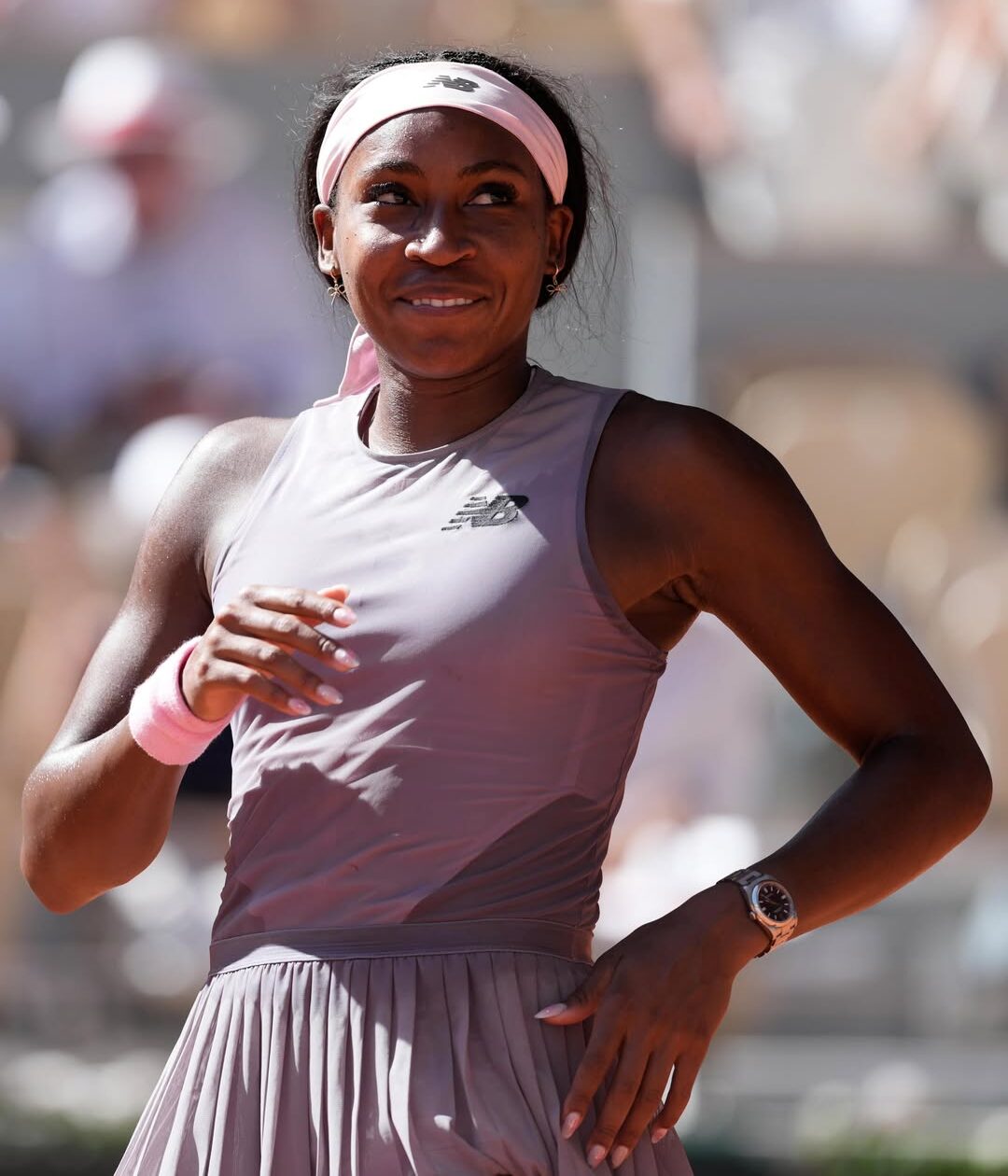 American tennis player Coco Gauff smiling on the clay court at Roland-Garros during her 2026 French Open tournament match, wearing a mauve New Balance kit and pink headband.
