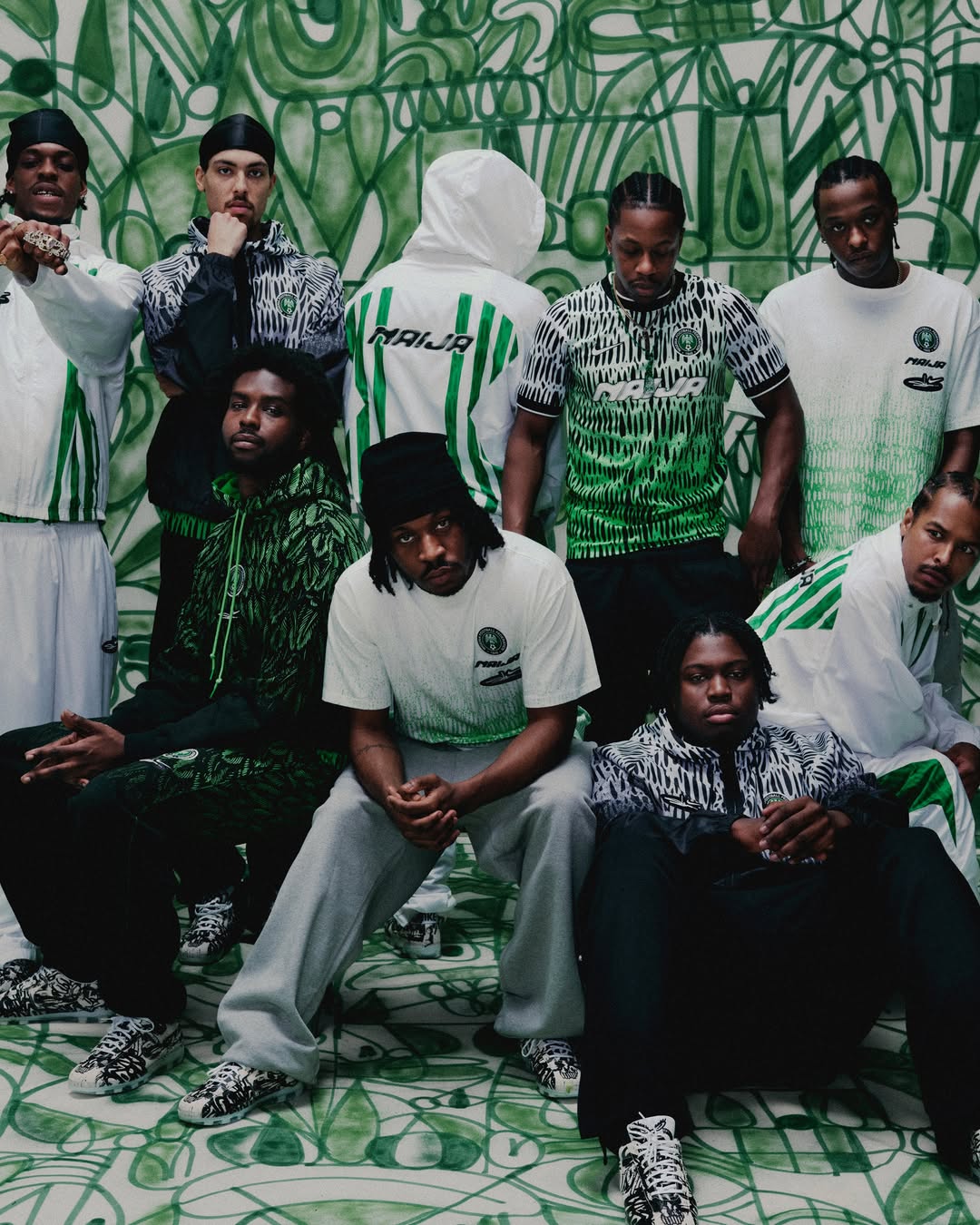 A group of models posing together against a green-and-white graffiti wall, showcasing various tracksuits, graphic jerseys, hoodies, and anthem jackets from the Nike x Slawn 2026 capsule collection.
