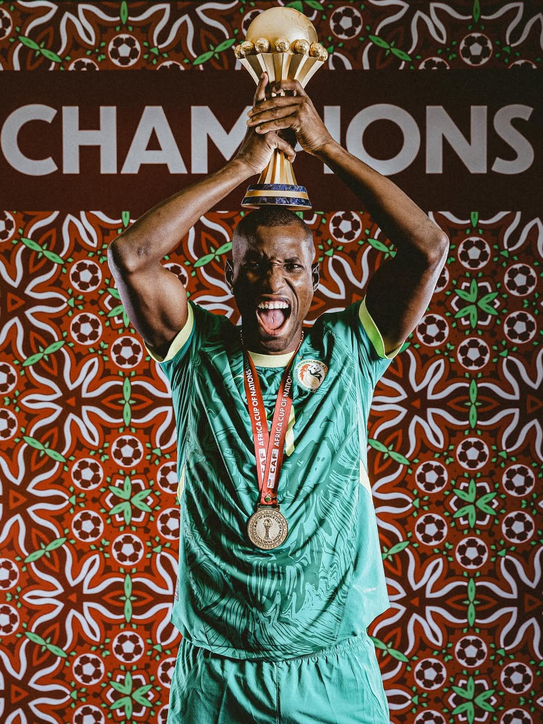 Senegal forward Nicolas Jackson screaming in celebration while holding the gold Africa Cup of Nations (AFCON) trophy over his head in front of a decorative "Champions" backdrop.