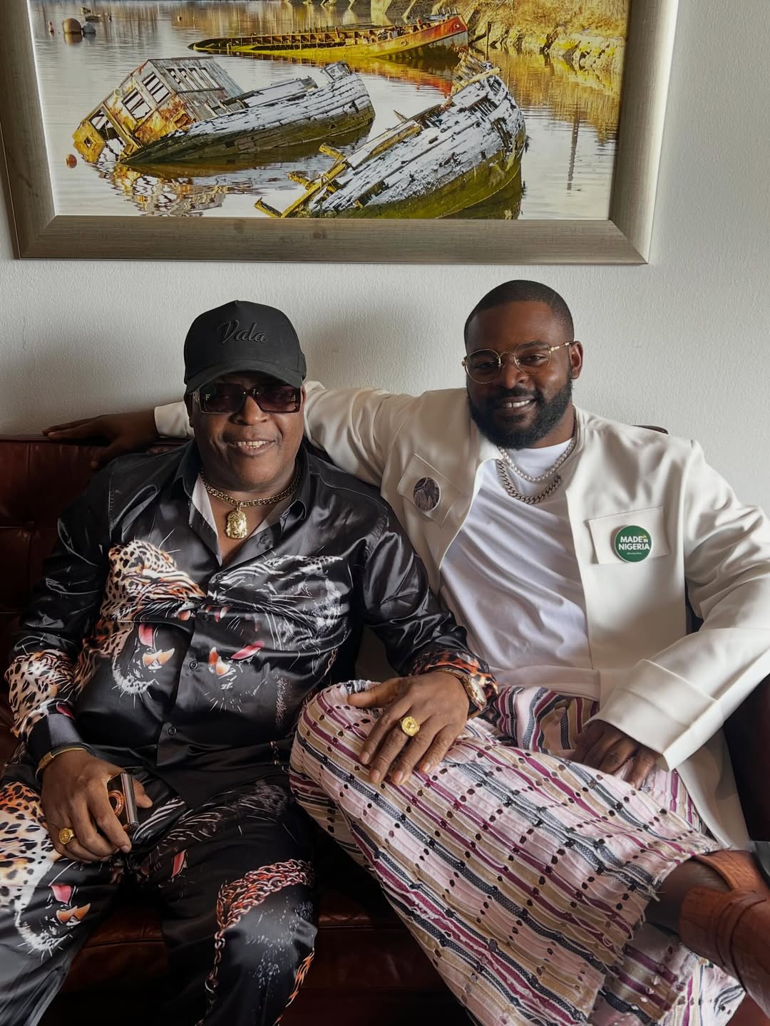 Legendary Juju musician Sir Shina Peters wearing a black graphic satin shirt alongside rapper Falz in a white jacket, sitting together on a leather sofa.