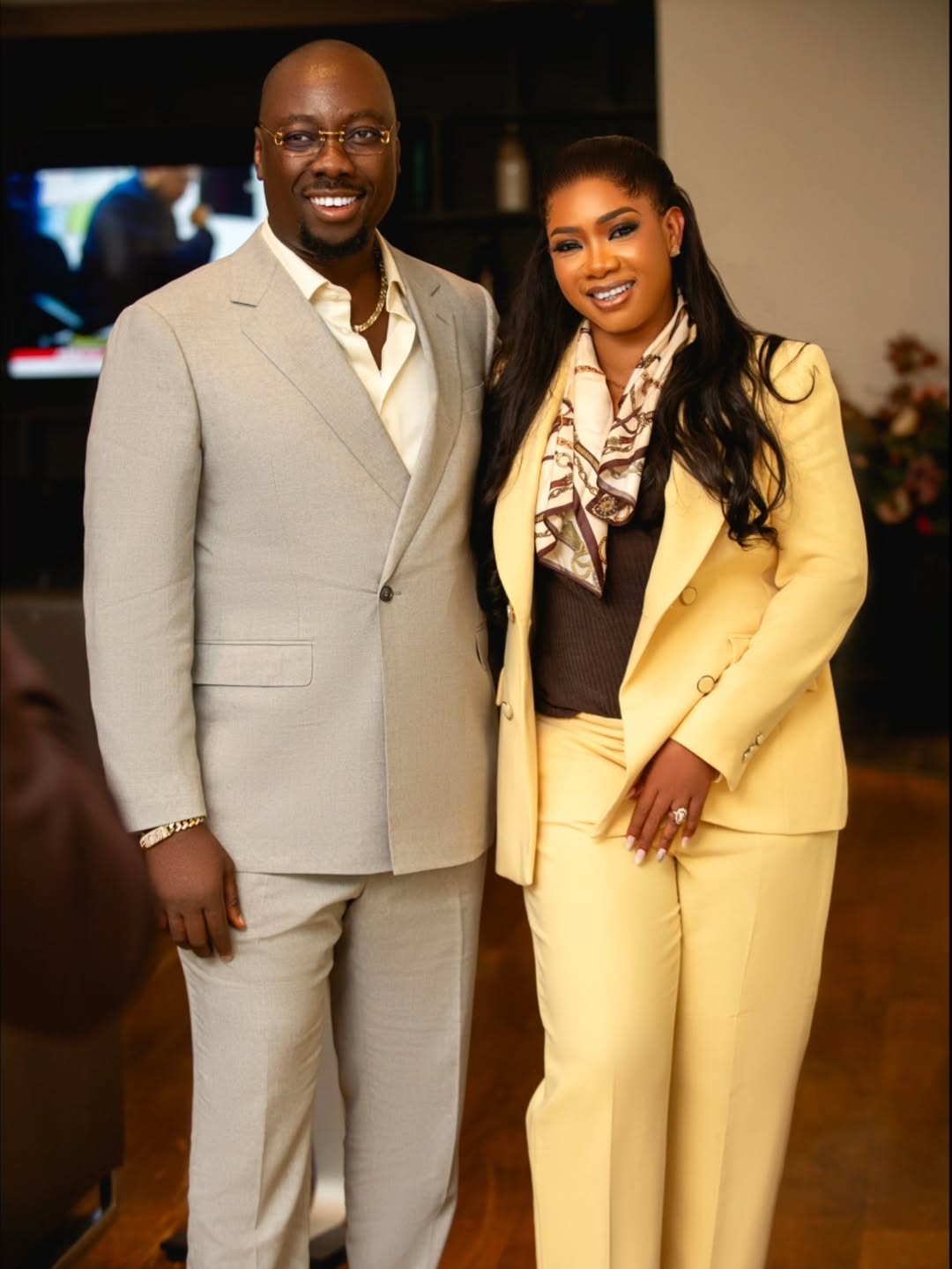 Full portrait of Nigerian entrepreneur Obi Cubana in a grey suit next to his wife Ebele Iyiegbu in a yellow double-breasted suit and silk scarf.